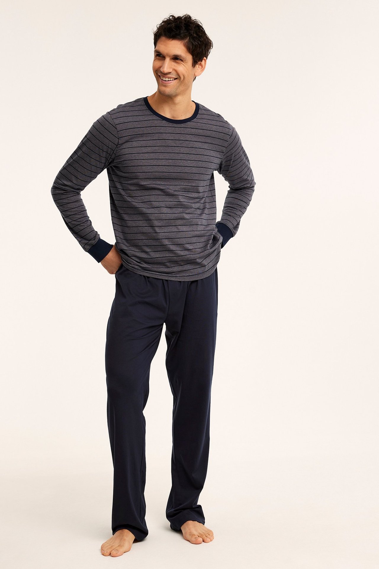 Miehen kokovartalokuva edestä: tummansiniset jersey-pyjamahousut ja harmaa raidallinen pitkähihainen paita.