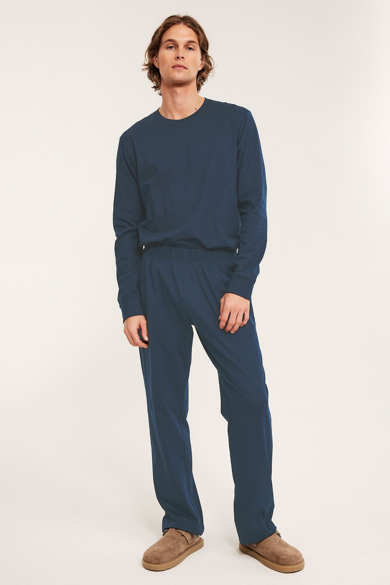 Mies edestä: tummansiniset puuvillapyjamahousut, yhteensopiva pitkähihainen paita ja ruskeat mokkanahkatossut.