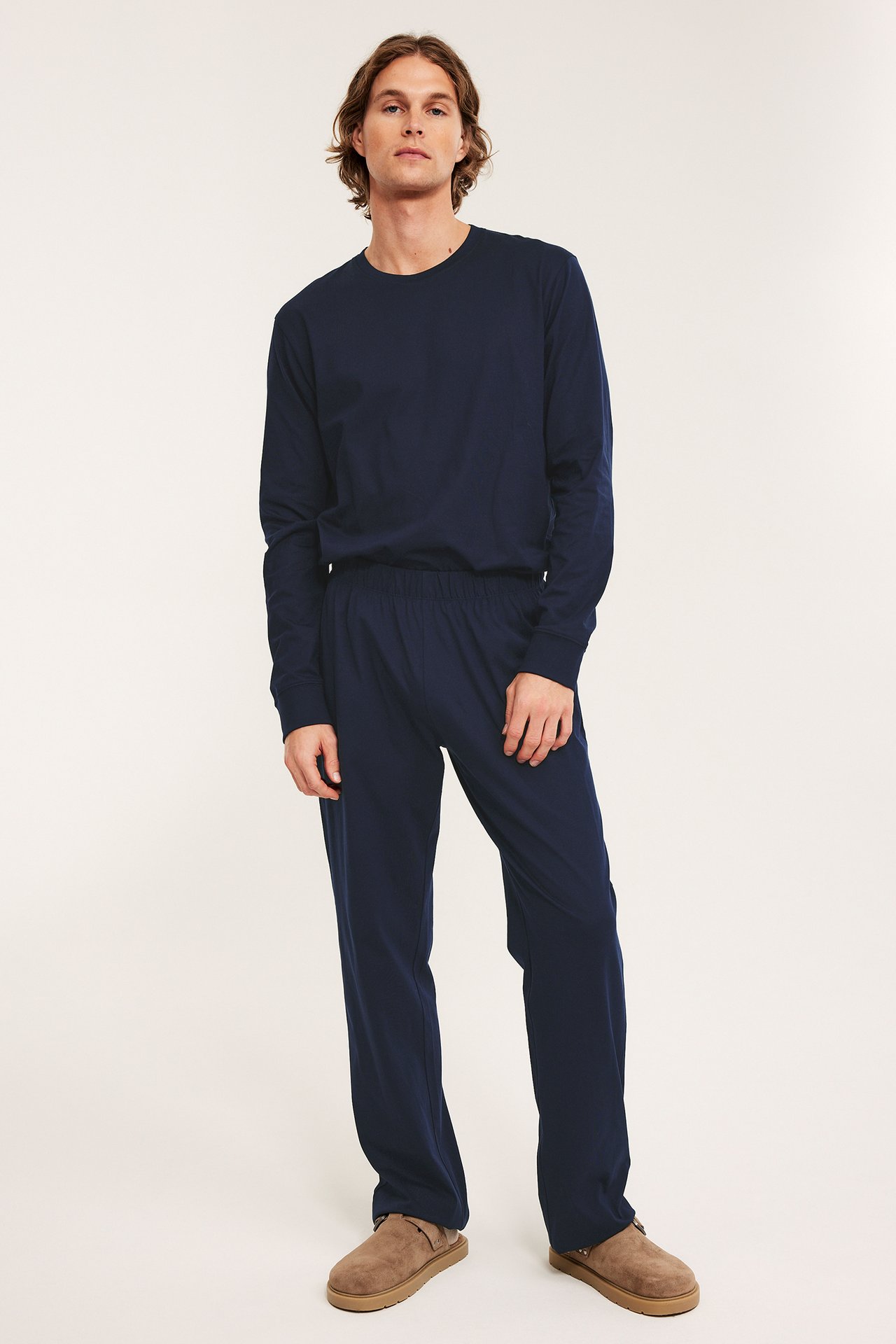 Mężczyzna z przodu w granatowych piżamowych spodniach z dżerseju, pasującej bluzce i brązowych kapciach.