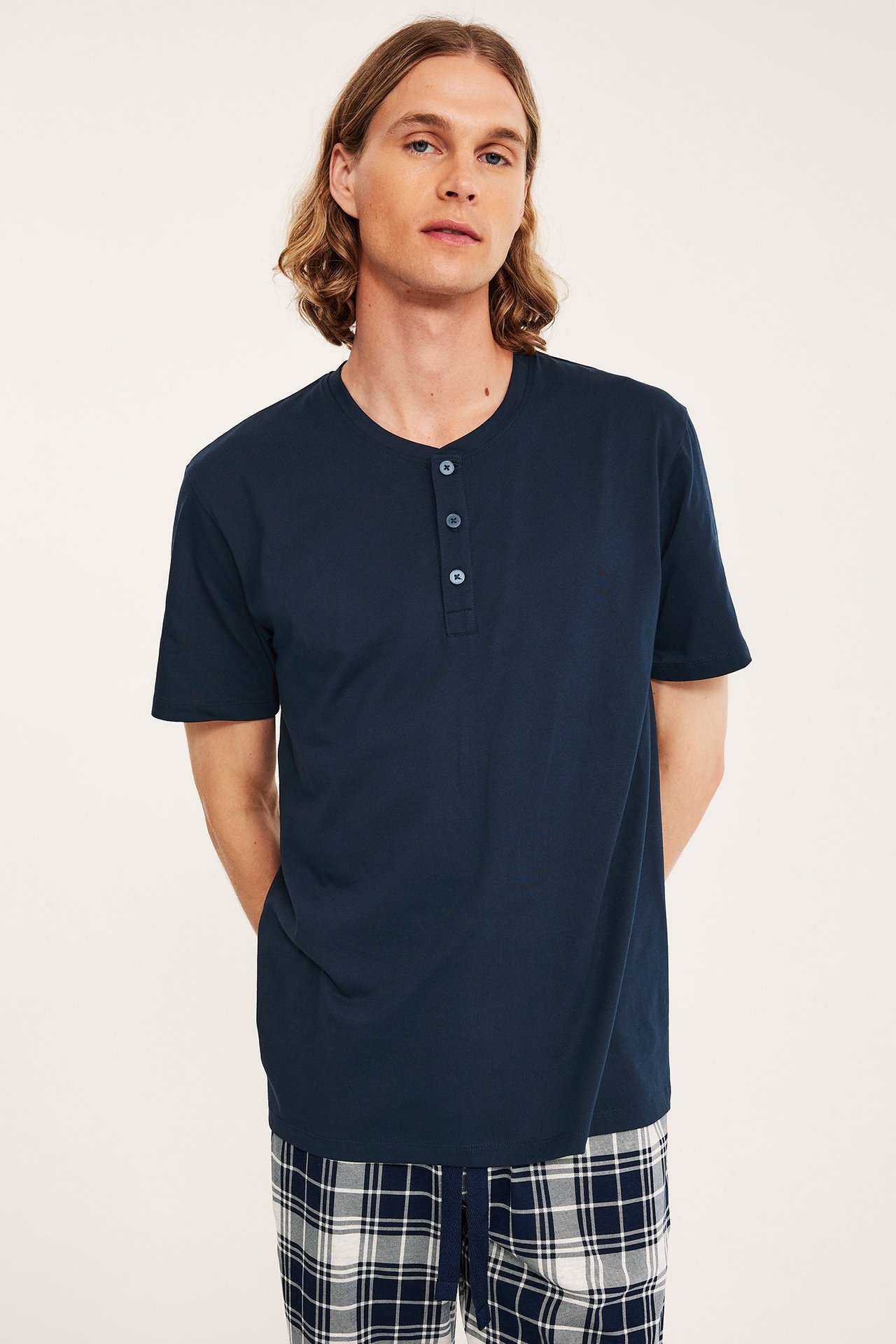 Framifrån: man i mörkblå kortärmad pyjamaströja i bomull med knappslå och rutiga pyjamasbyxor.