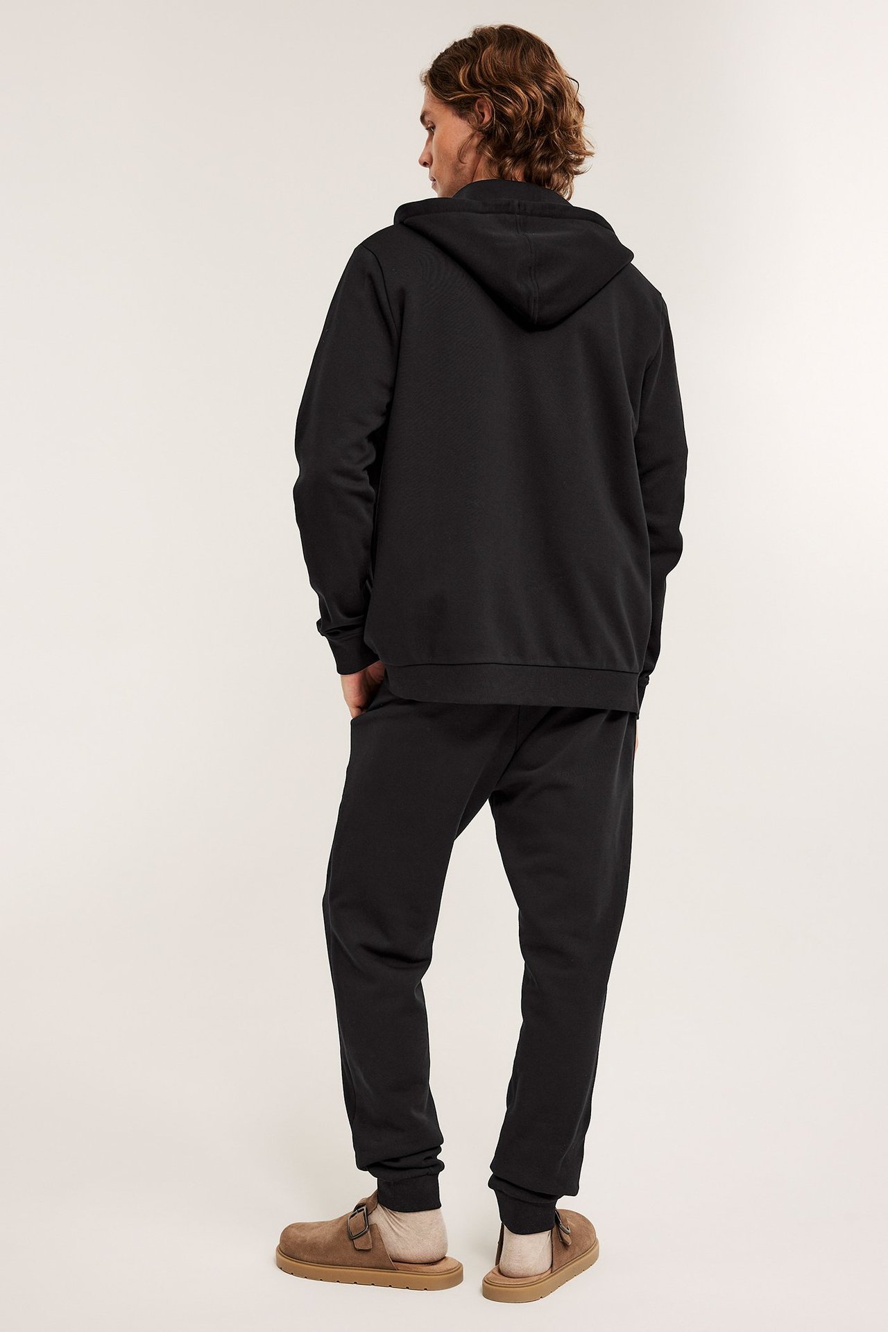 Mies takaapäin mustissa collegehousuissa, joissa resorit, musta huppari ja vaaleanruskeat tohvelit.