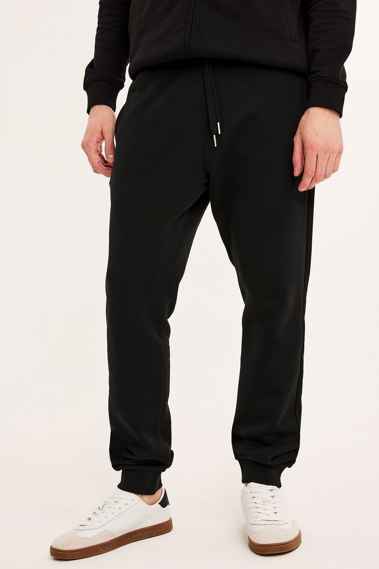 Framifrån, svarta herrsvettisbyxor med ribbade muddar, burna med svart huvtröja och vita sneakers.