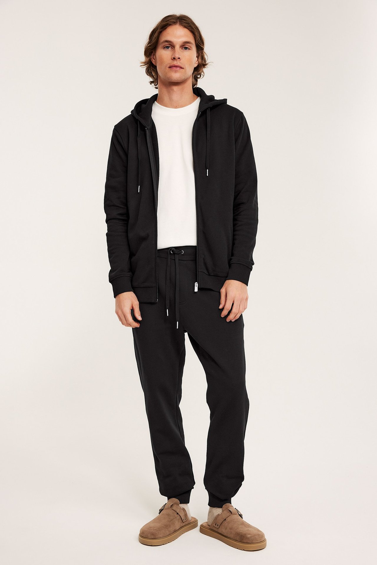 Mies mustissa kiristysnyörillisissä collegehousuissa, musta vetoketjuhuppari, valkoinen t-paita ja ruskeat tohvelit.