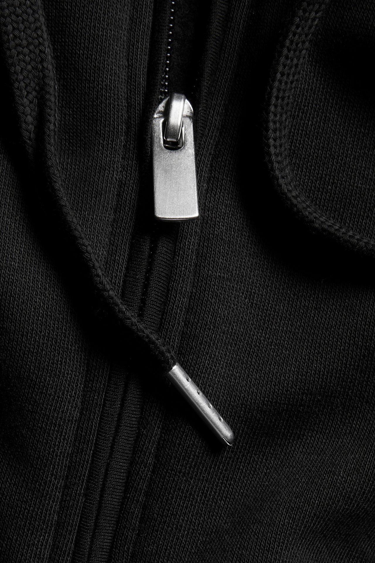 Zbliżenie na czarną bluzę z kapturem na zamek, ukazujące metalowy suwak i sznurek z metalową końcówką.
