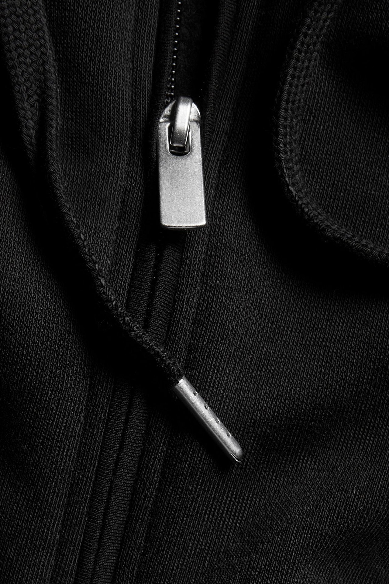 Närbild av en svart huvtröja med dragkedja, visar metallhandtaget och en dragsko med metallspets.