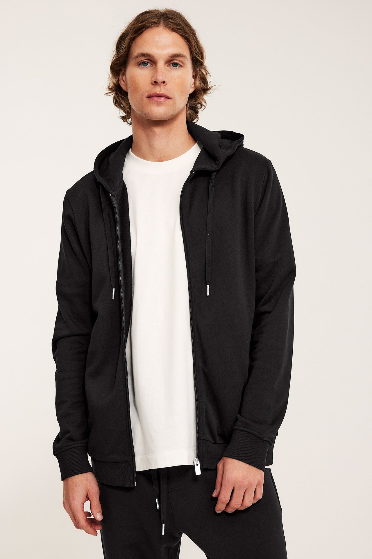 Widok z przodu mężczyzny w czarnej rozpinanej bluzie z kapturem, białym T-shircie i czarnych spodniach.