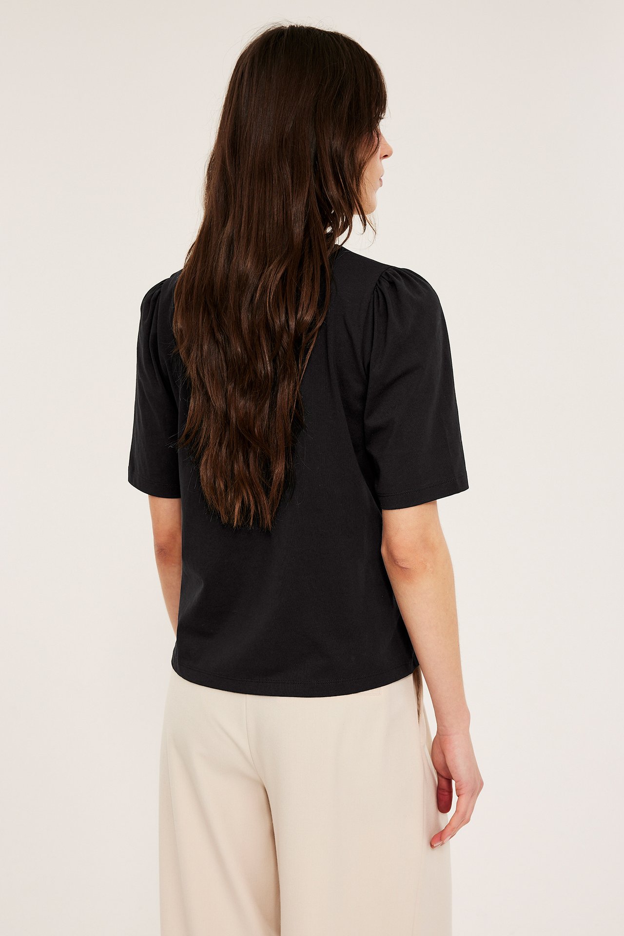 Bakifrån: kvinna i svart bomullstopp med puffärm och ljusbeige byxor.