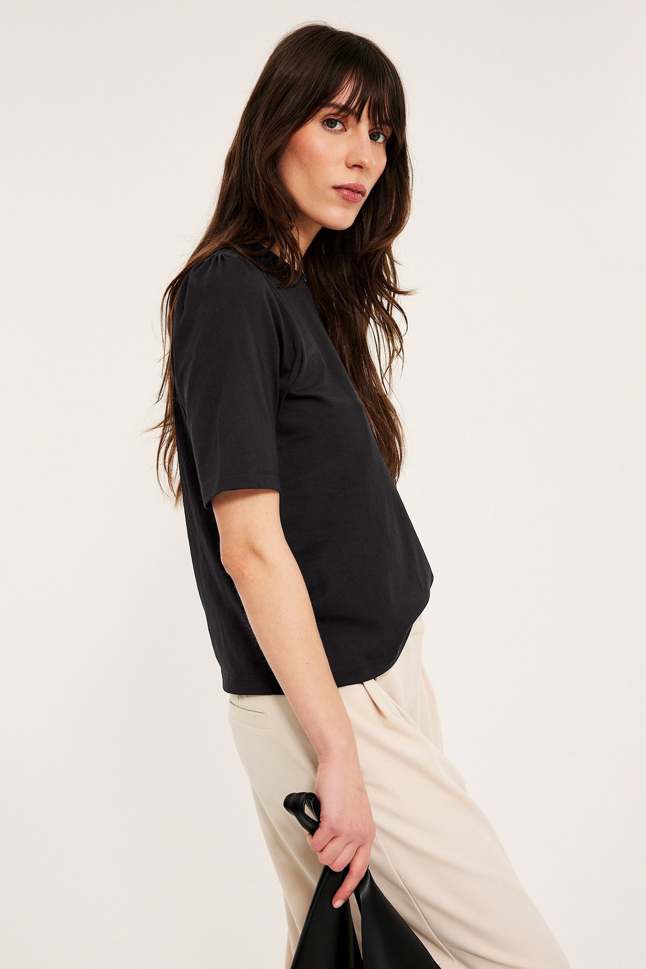 Sidovy av kvinna i svart bomullstopp med puffärm, ljusbeige byxor och med en svart handväska.