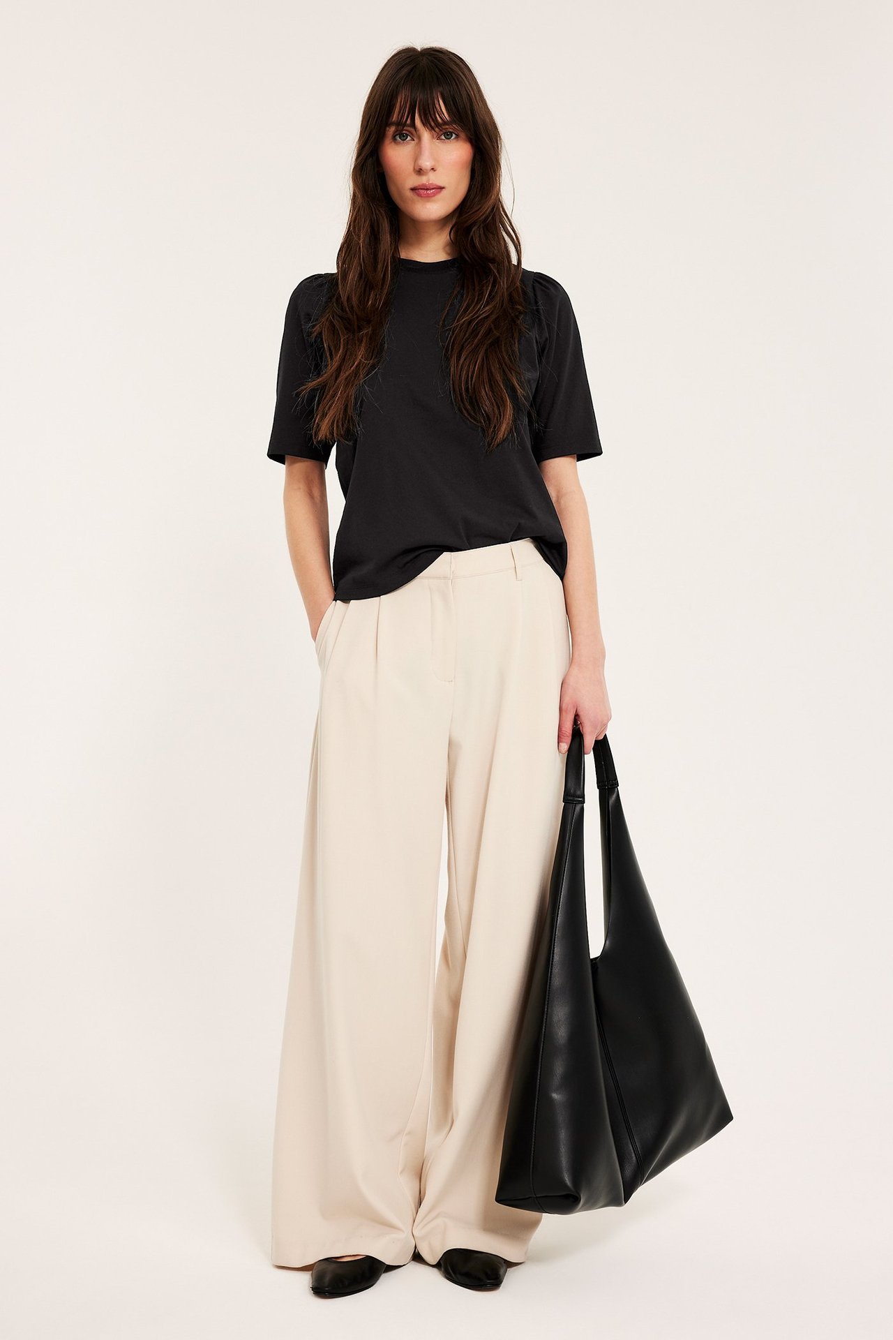 Kobieta ma czarny top z bufiastymi rękawami, beżowe spodnie, czarne płaskie buty i czarną torebkę.