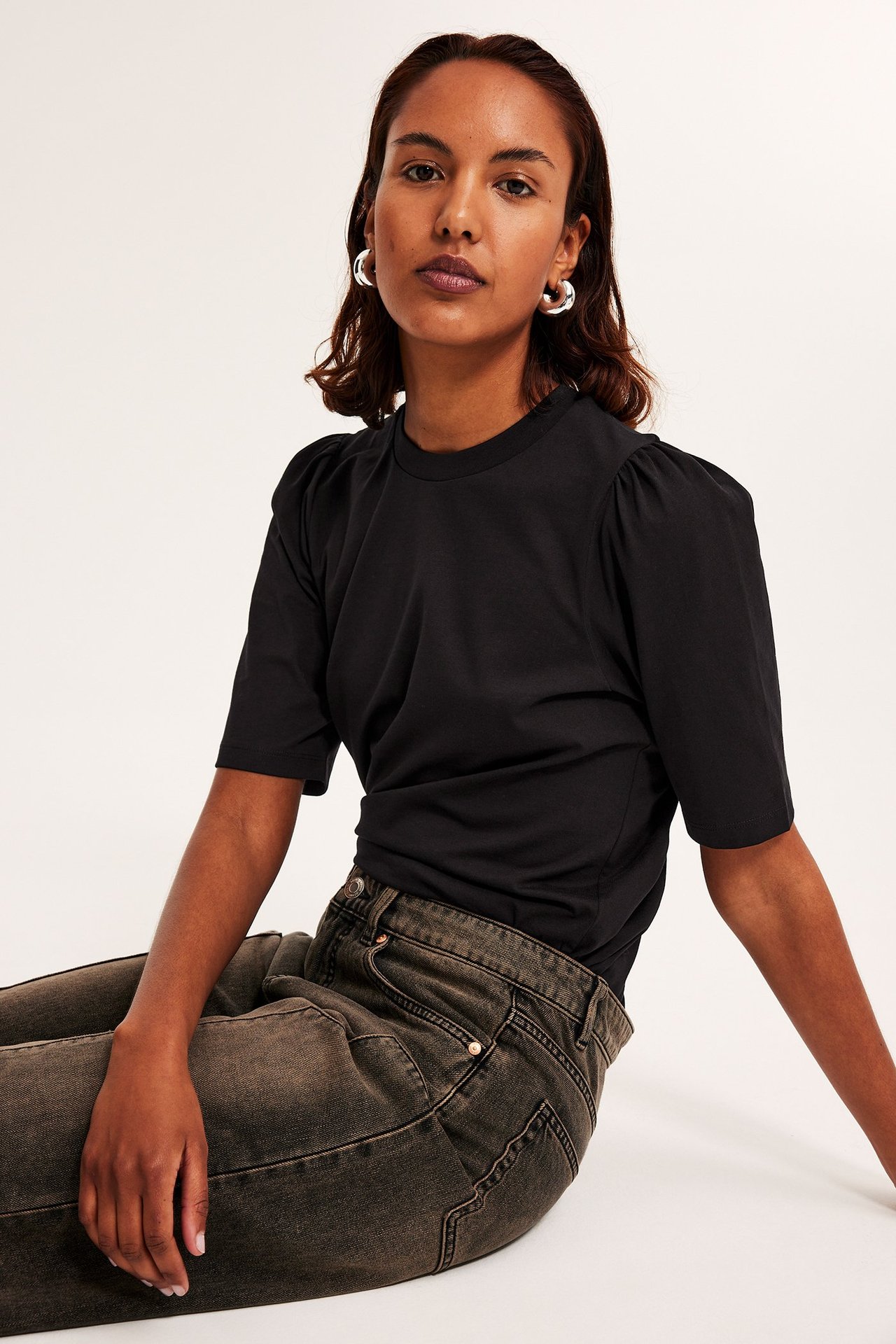 Frontbilde av en kvinne i svart bomullstopp med puffermer, mørkegrå jeans og sølvfargede øreringer.