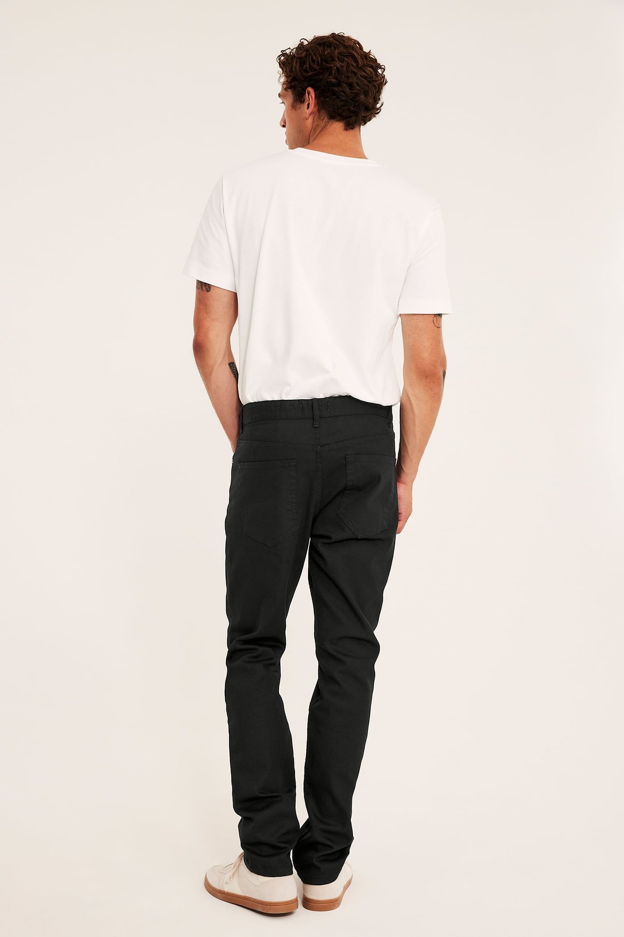 Bakfra en mann i sorte rette bukser, hvit t-skjorte og beige joggesko.