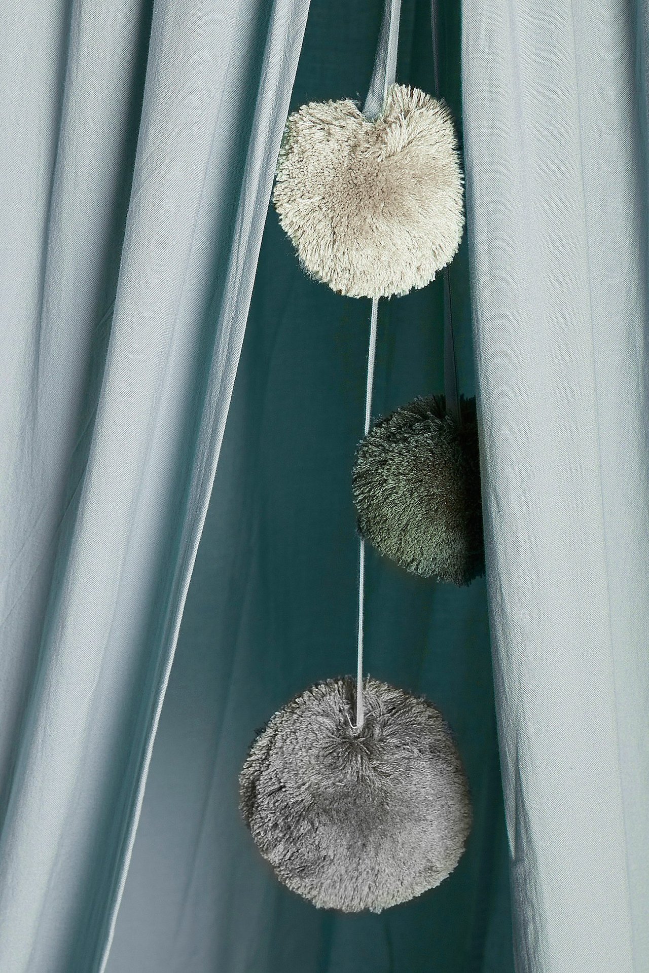 Zbliżenie jasnoniebieskiego namiotu do czytania z trzema wiszącymi pomponami w beżu, ciemnej zieleni i szarości.