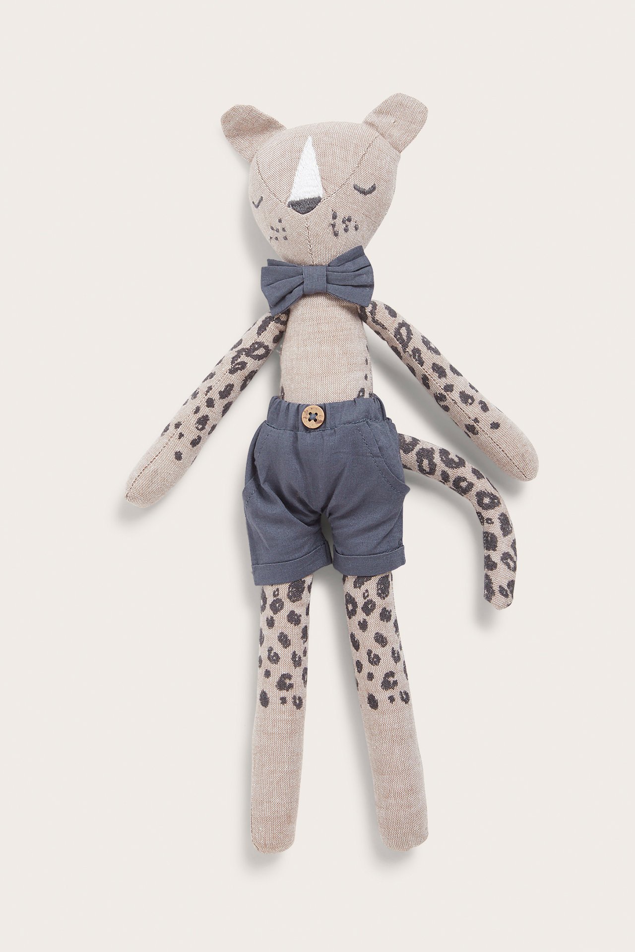 Edestä näkymä beigestä leopardipehmolelusta, jossa tummanharmaita täpliä, yllään harmaat shortsit ja rusetti.