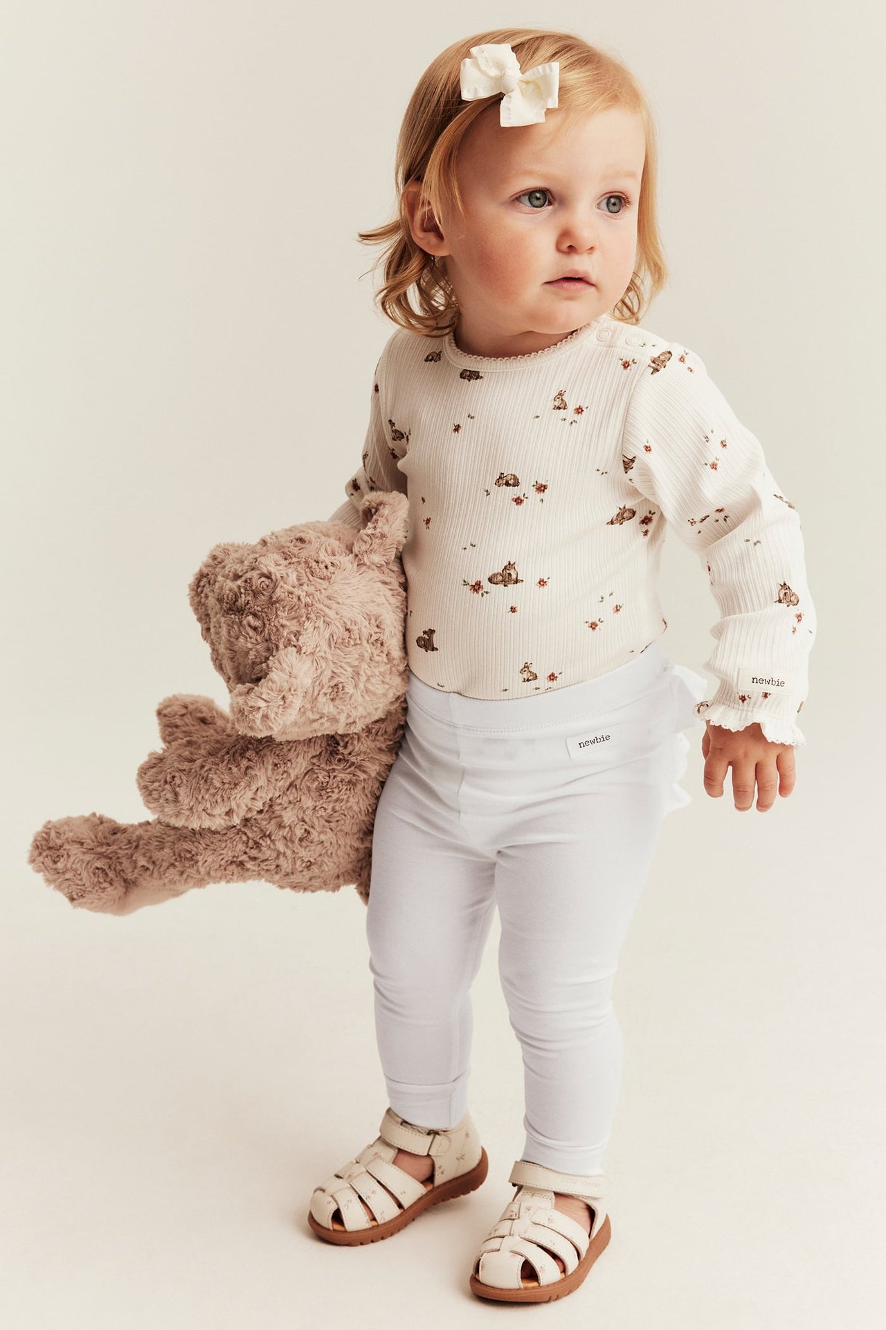 Vauva valkoinen peura-kuviollinen body, valkoiset leggingsit ja sandaalit, pitää nallea.