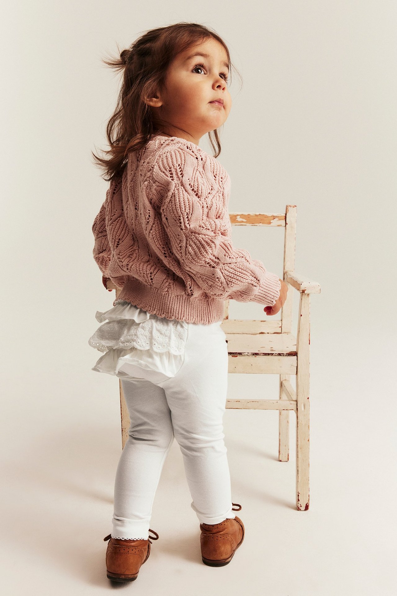 Sned bakvy av baby i offwhite leggings med volang, rosa stickad tröja och bruna skor.