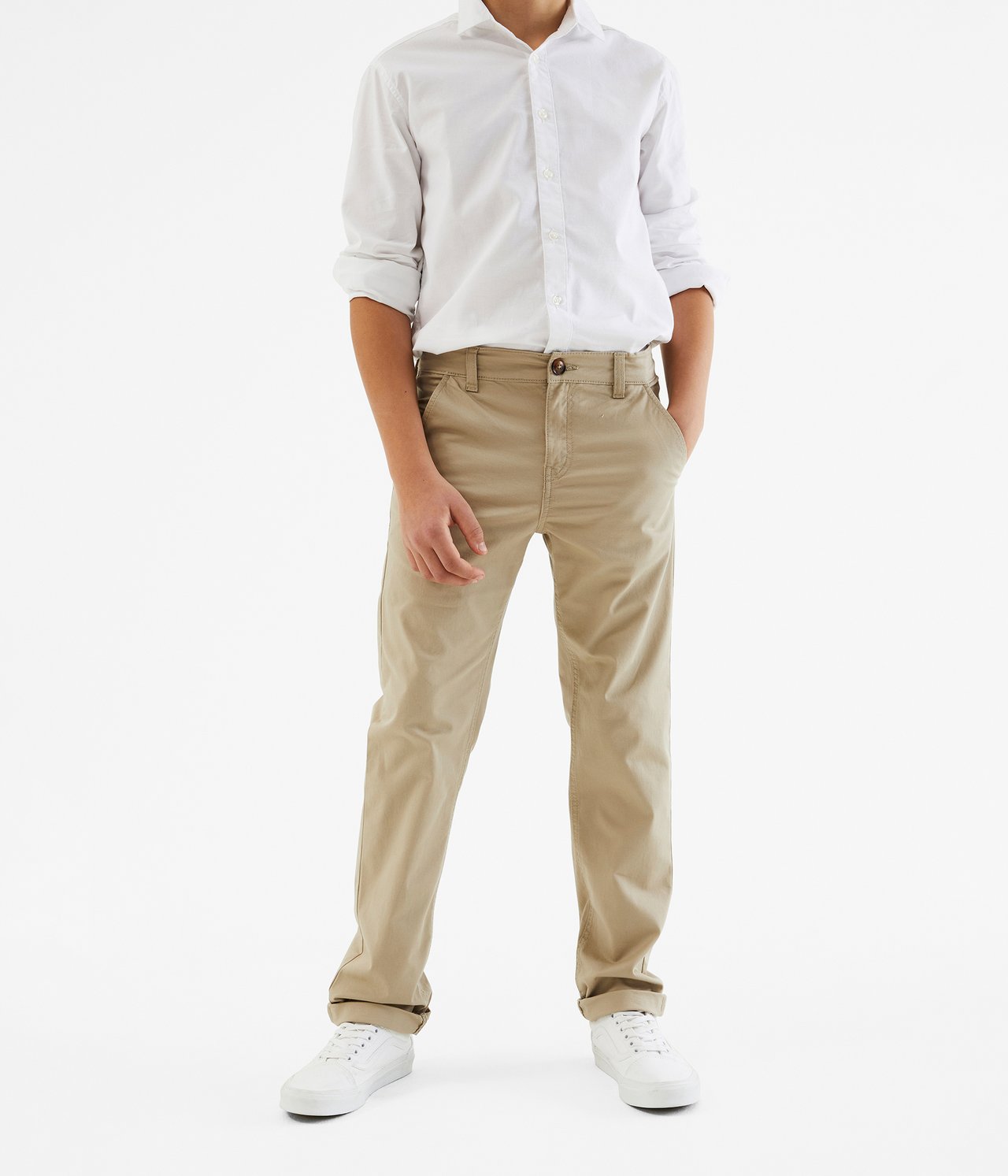 Frontvy av ett barn som bär beiga stretchchinos med uppvikta ben, en vit långärmad skjorta och vita sneakers.