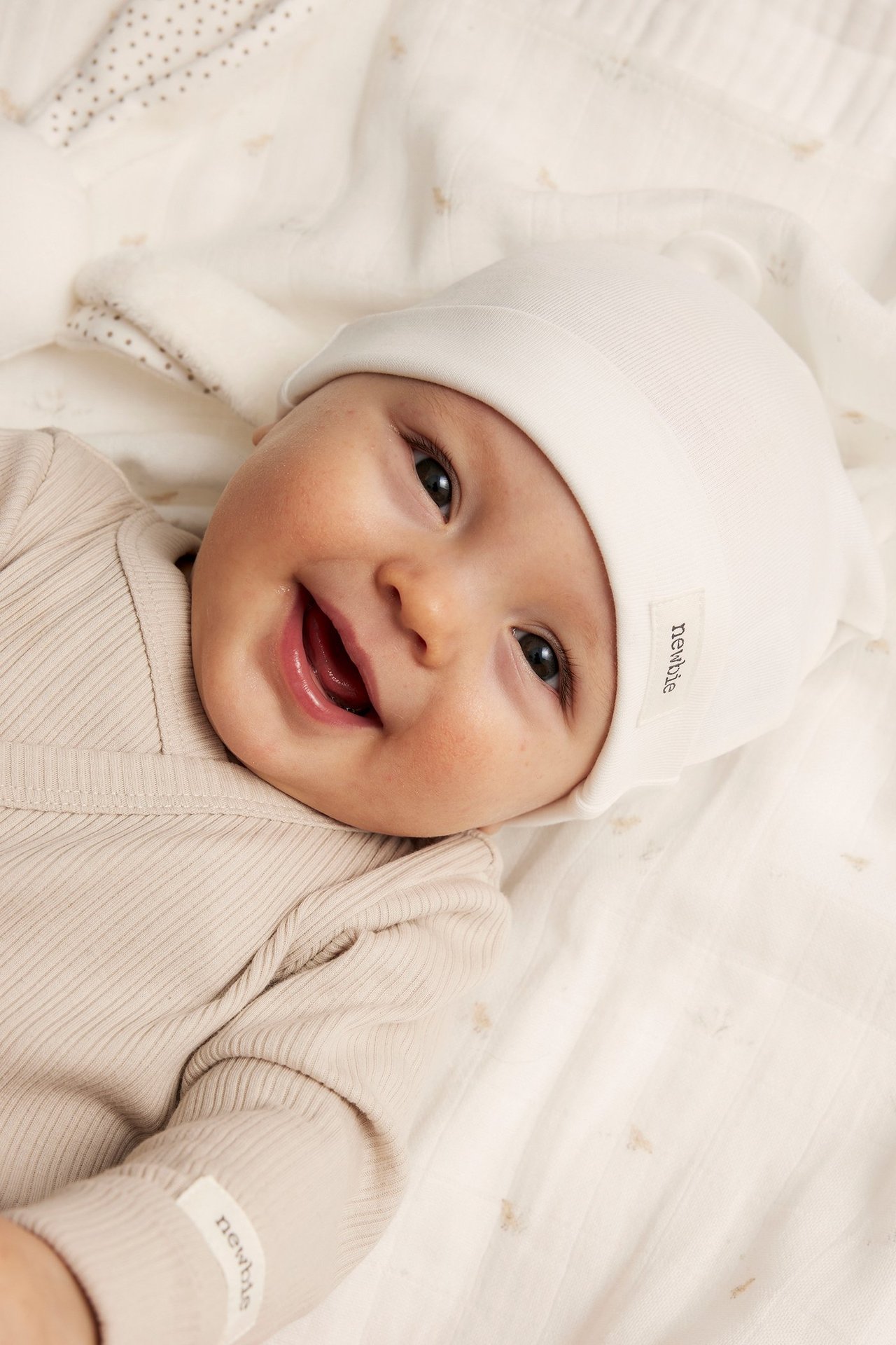 Närbild på en leende baby i en offwhite bomulls-trikåmössa och en ljusbeige ribbad body.