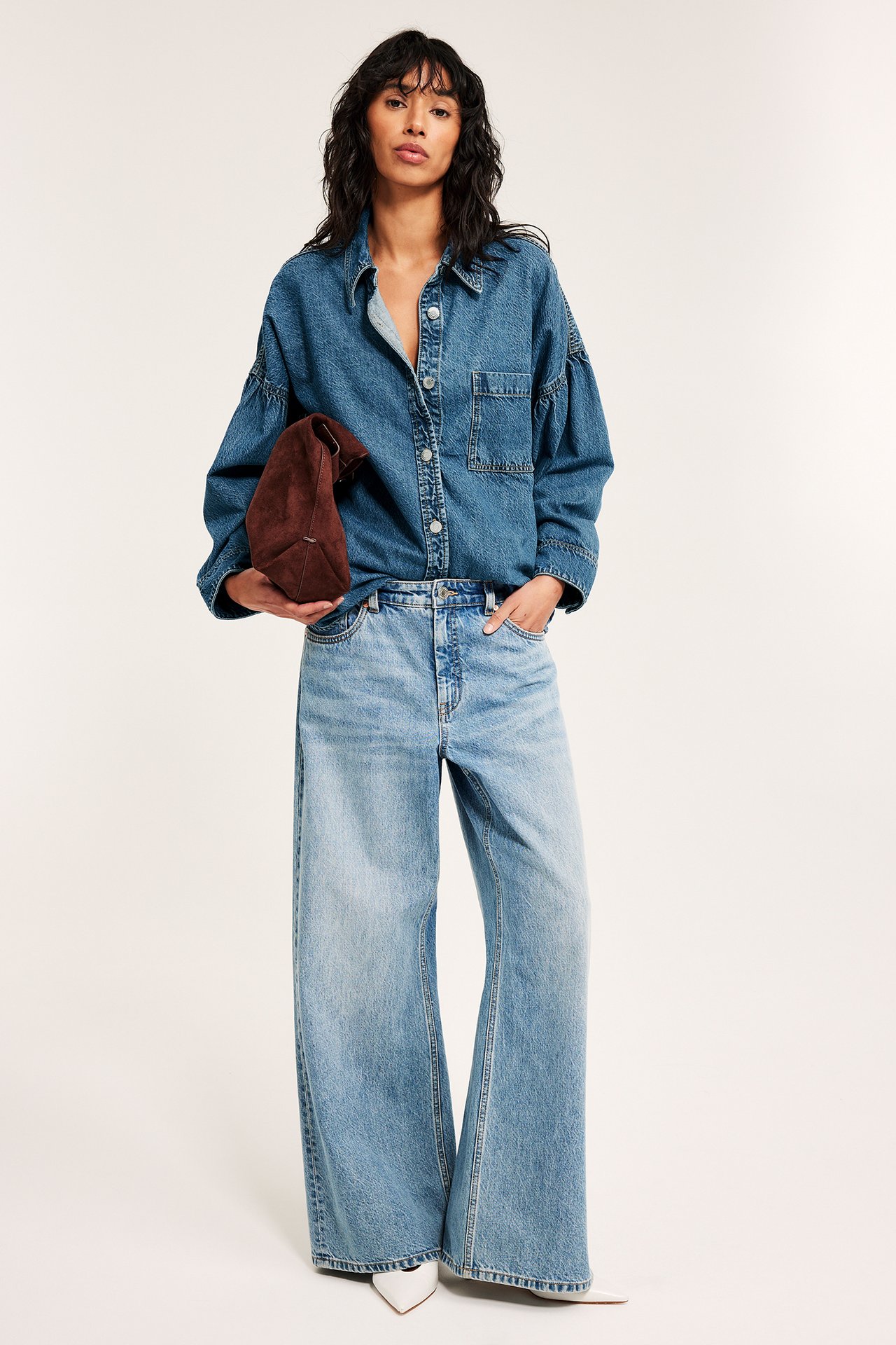 Edestä, nainen yllään sininen denim-paita drapeeratuilla hihoilla, vaaleat leveälahkeiset farkut, pitää ruskeaa käsilaukkua.