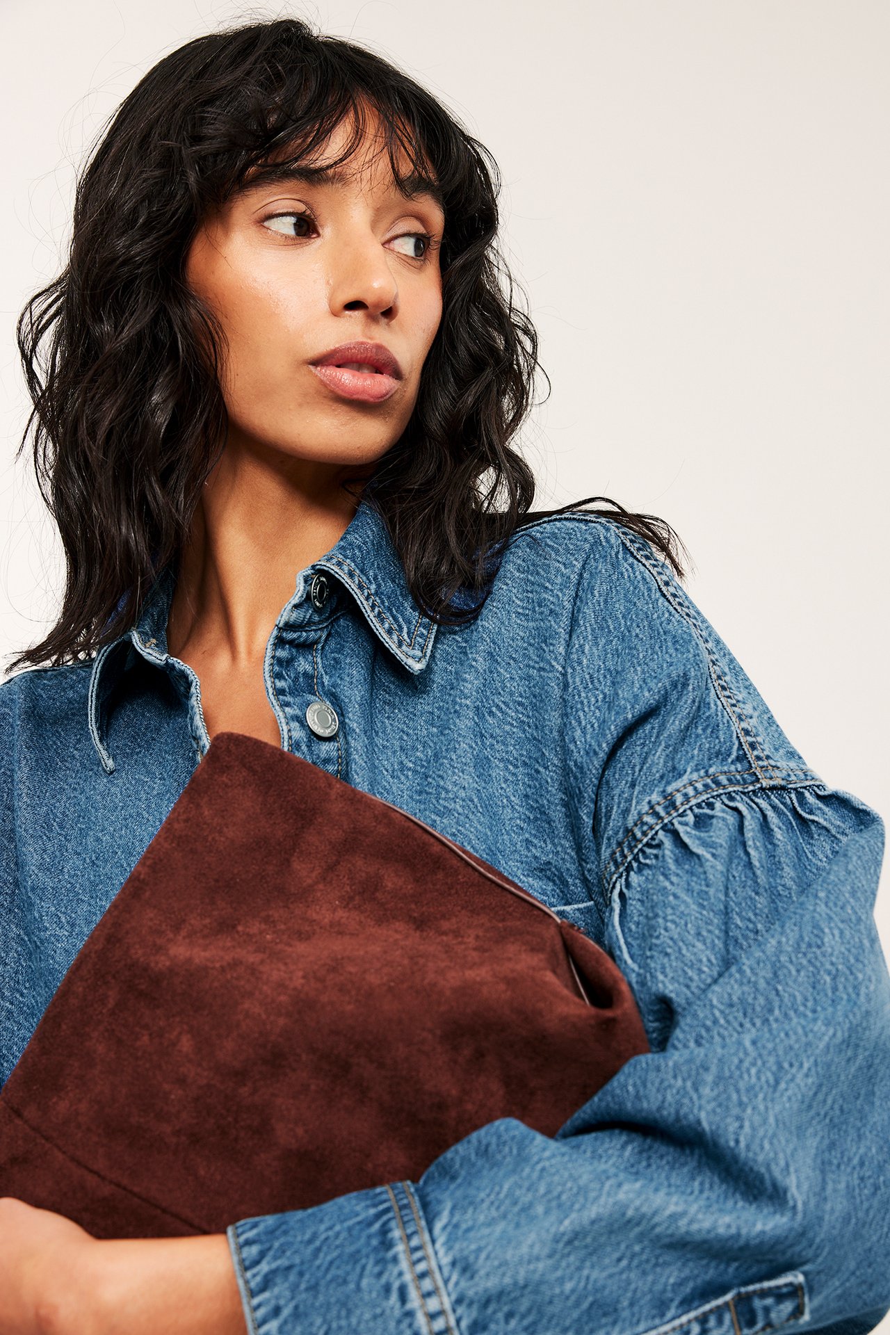 Lähikuva naisesta sinisessä denimpaidassa, jossa on drapeeratut hihat, pitäen tummanruskeaa clutch-laukkua.