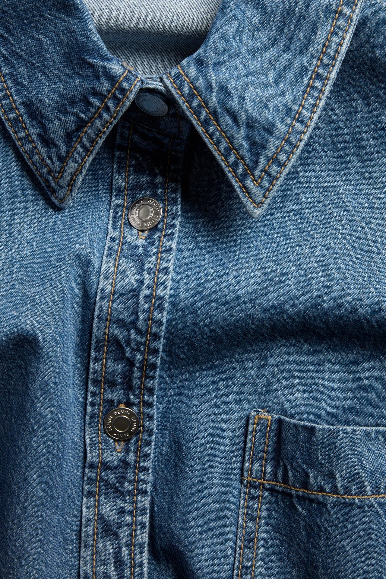 Zbliżenie z przodu na niebieską koszulę jeansową z kołnierzem, metalowymi napami i kieszenią.