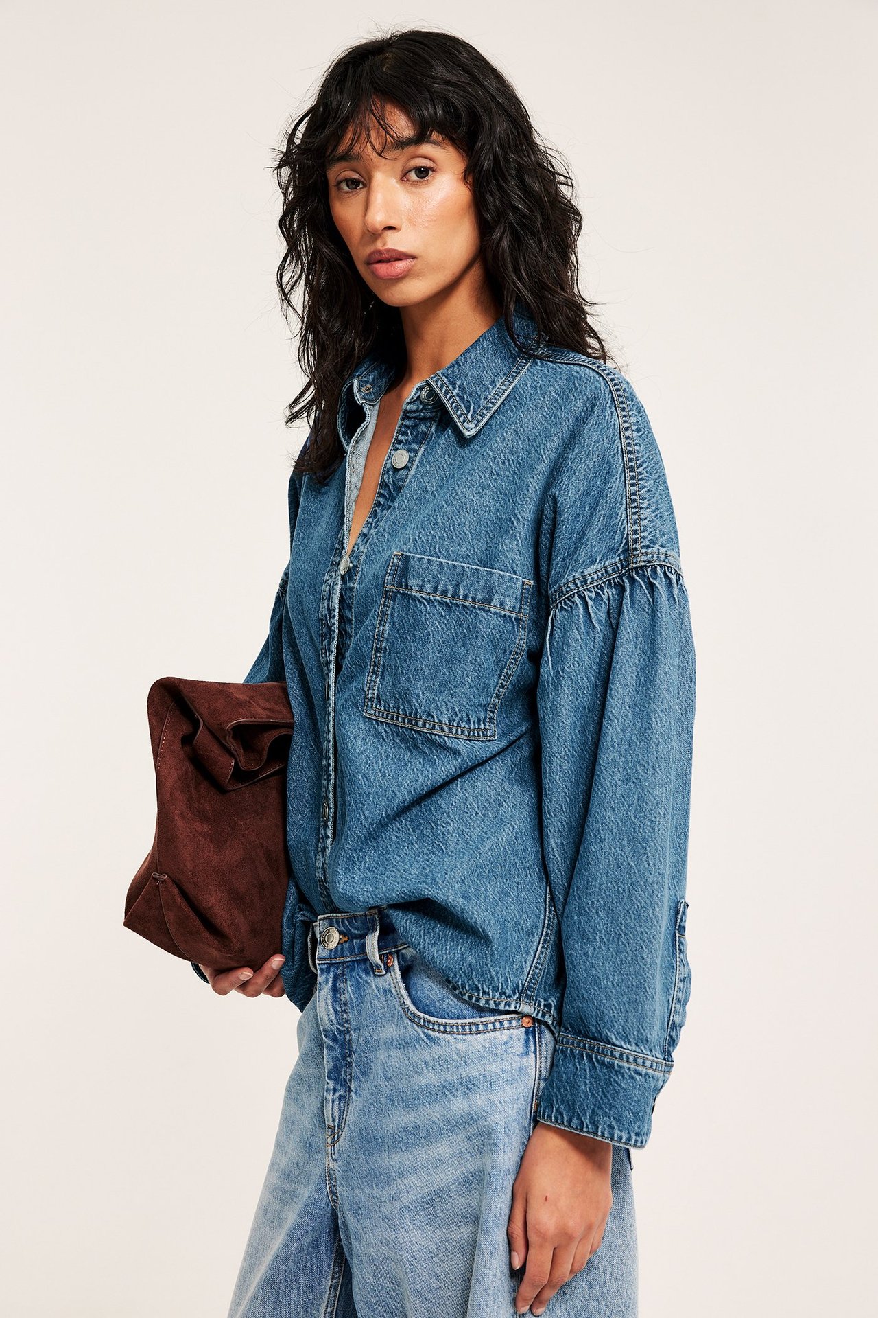 Naisen etukuva sinisessä denimpaidassa, laskostetut hihat, vaaleansiniset farkut, ruskea laukku.