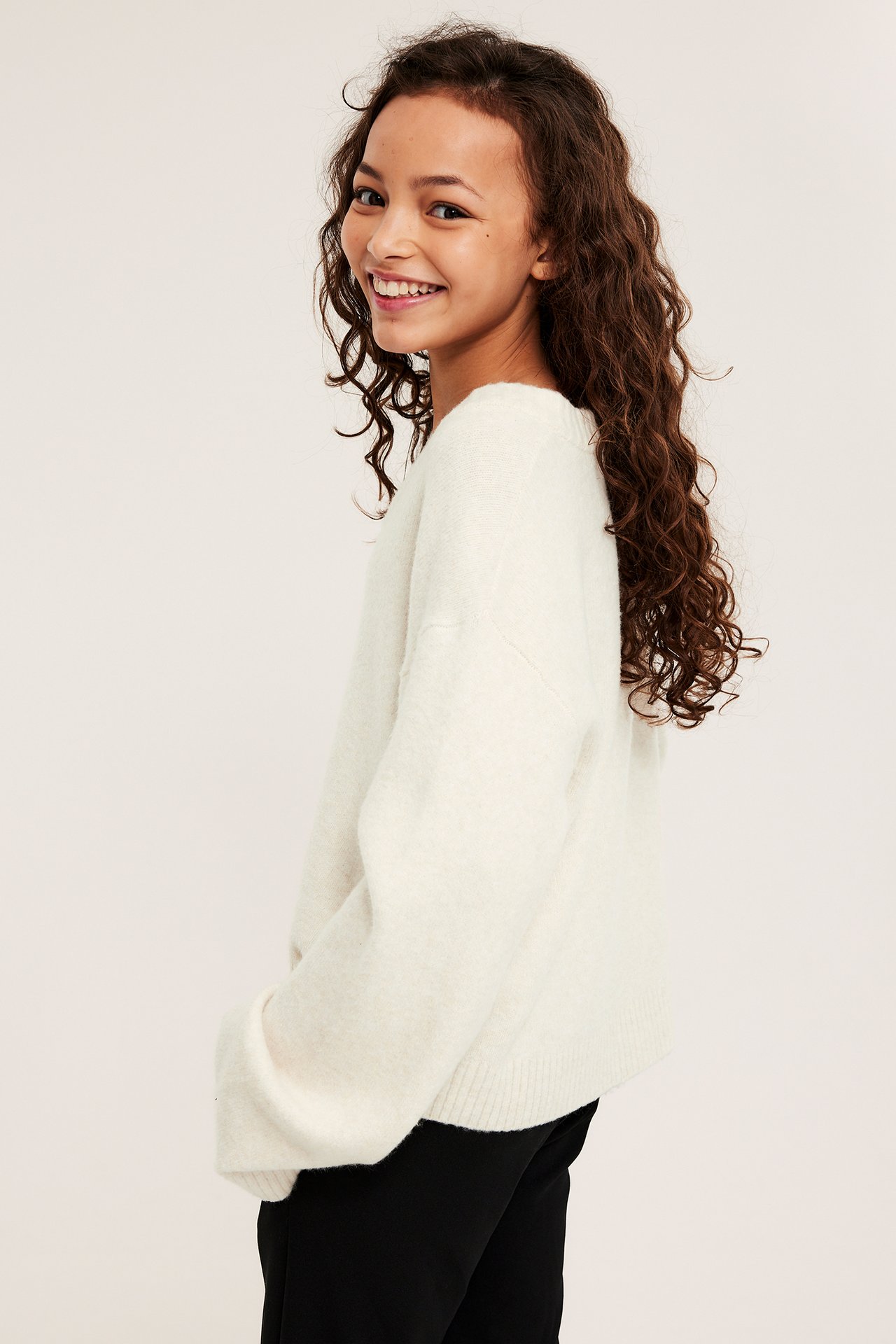 Widok z boku dziecka w białym melanżowym swetrze z dekoltem w serek na czarnych spodniach.