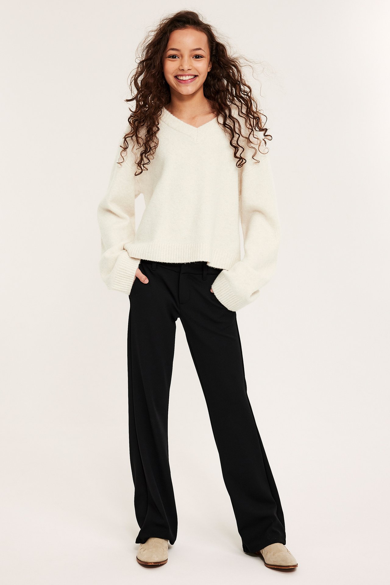 Widok z przodu: dziecko w kremowym melanżowym swetrze w serek, czarnych szerokich spodniach i jasnobeżowych butach.