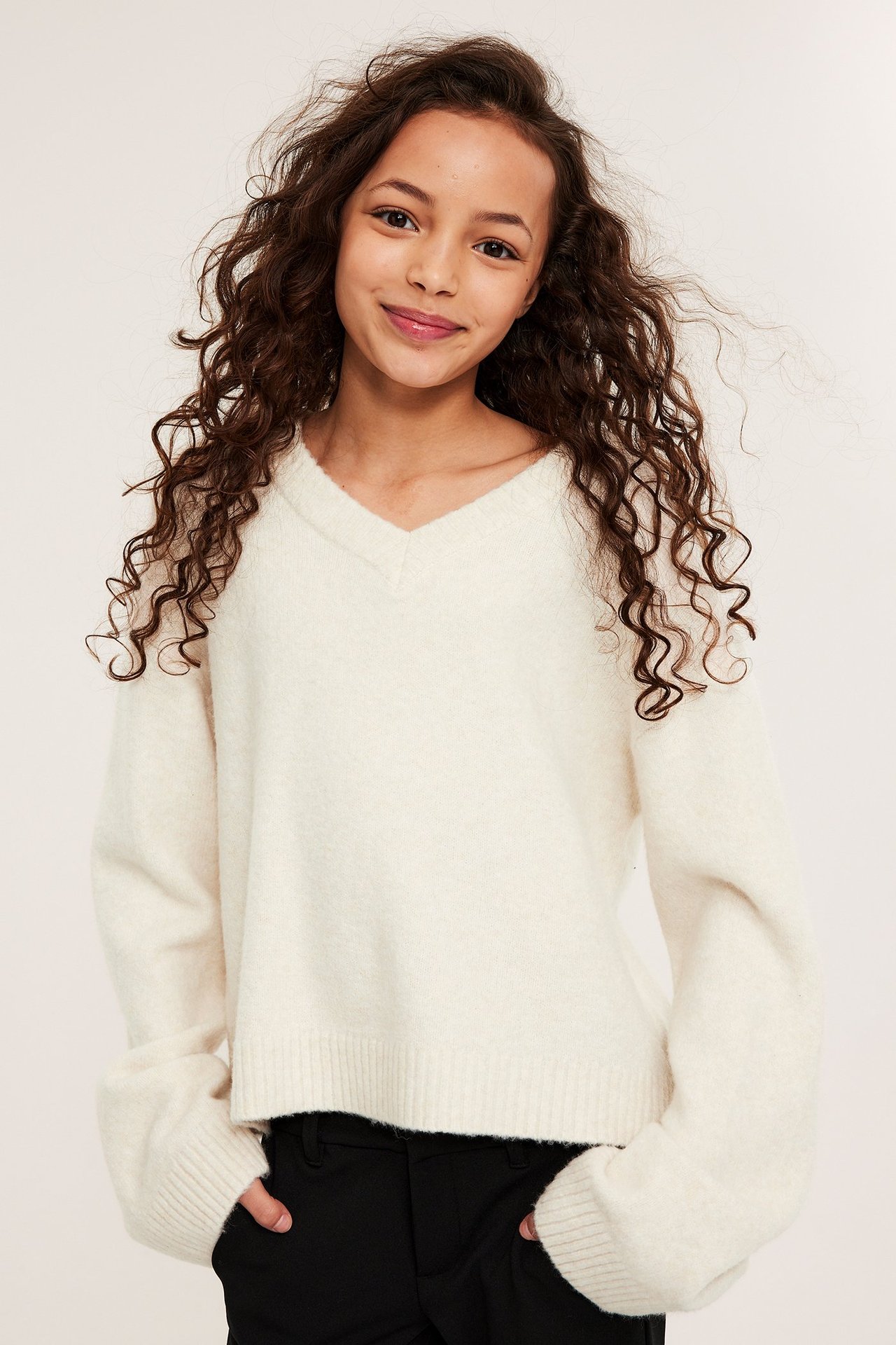 Frontvy av ett barn i en melerad vit v-ringad stickad tröja och svarta byxor.