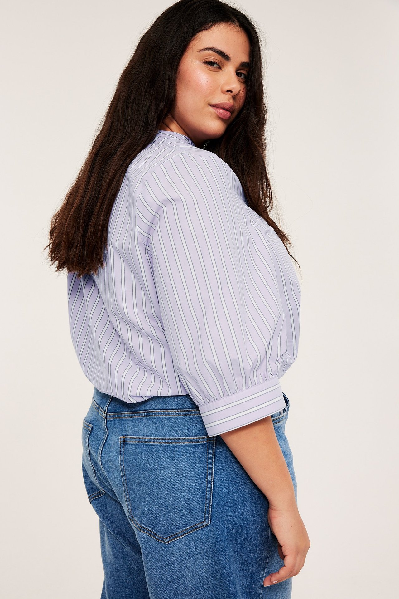 Nainen takaa ja sivulta yllään vaaleanliila raidallinen popliinipusero 3/4-hihoilla ja siniset farkut.
