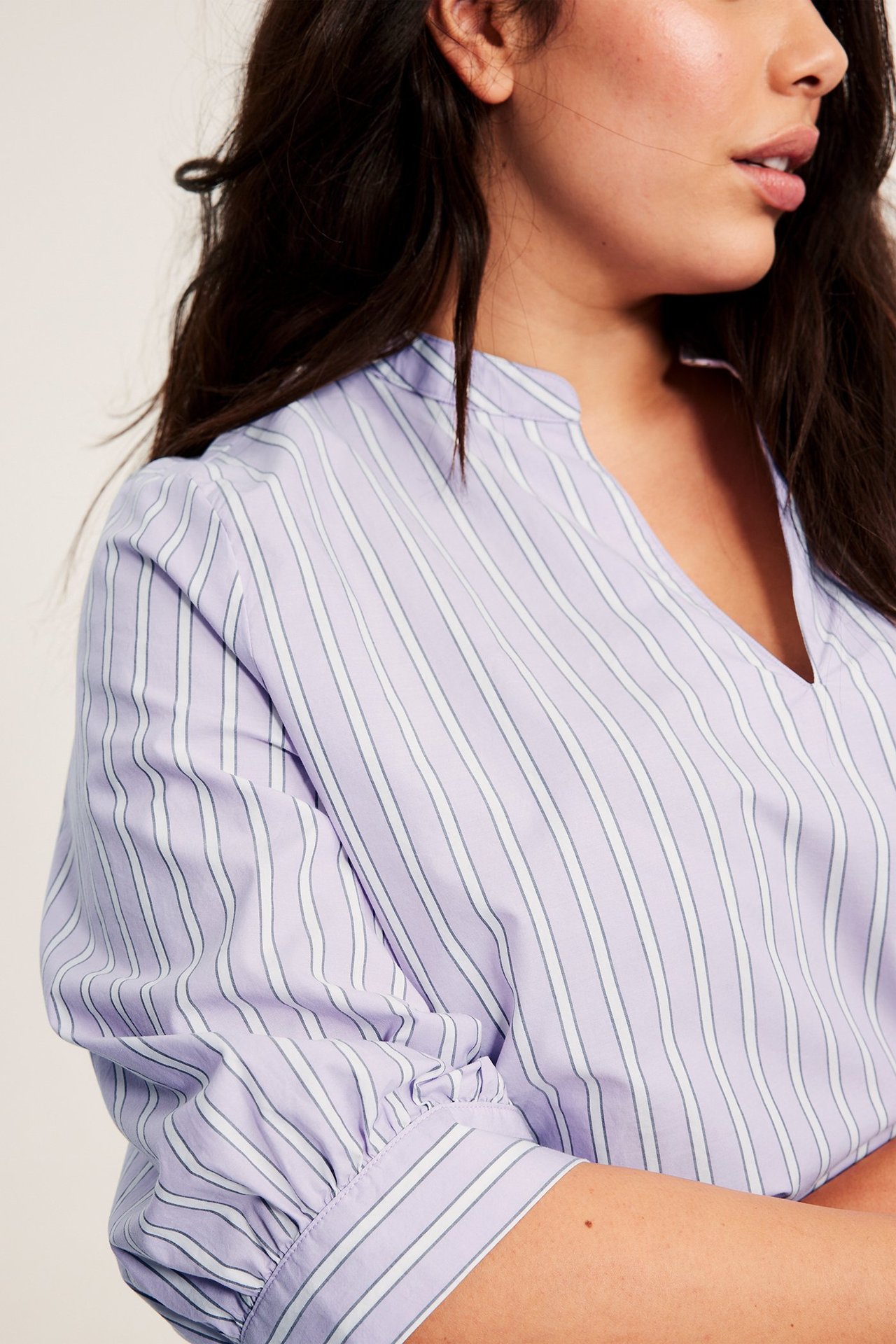 Lähikuva naisesta yllään vaaleanliila raidallinen popliinipusero, jossa V-aukko ja 3/4-pussihihat.