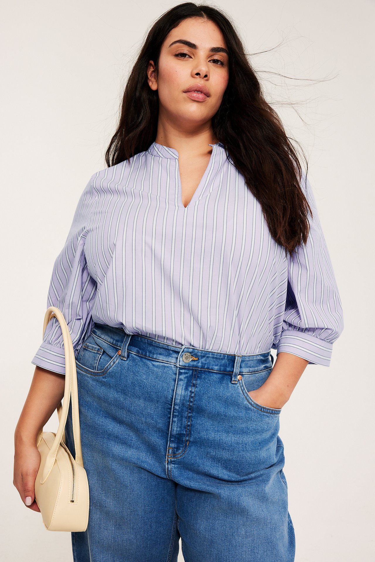 Framifrån: Kvinna i ljuslila randig poplinblus med 3/4-ärm, blå jeans, håller en krämfärgad handväska.