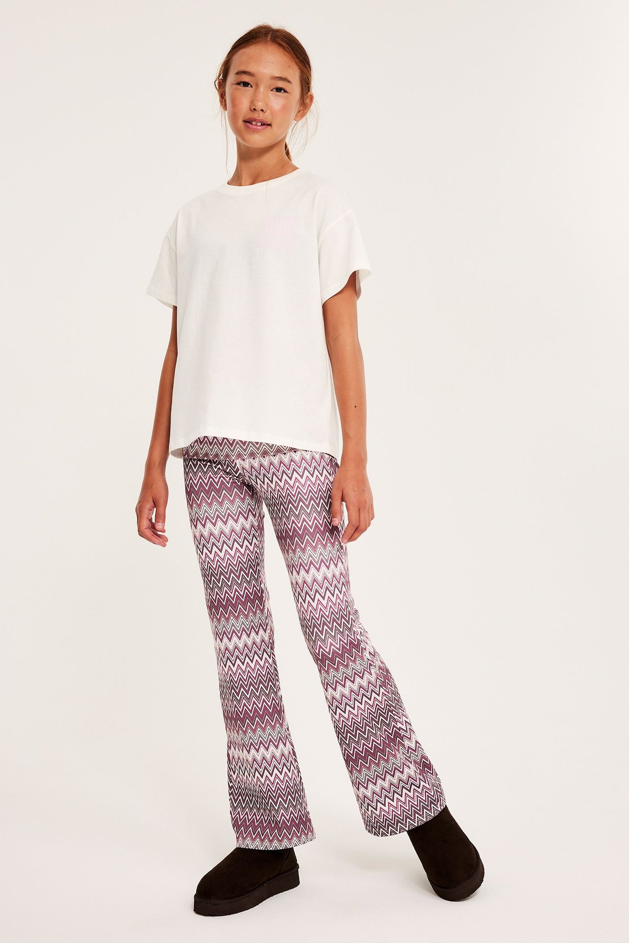 Edestä kuvattu lapsi yllään tummanpunaiset kuviolliset levenevät leggingsit, valkoinen t-paita ja tummat kengät.