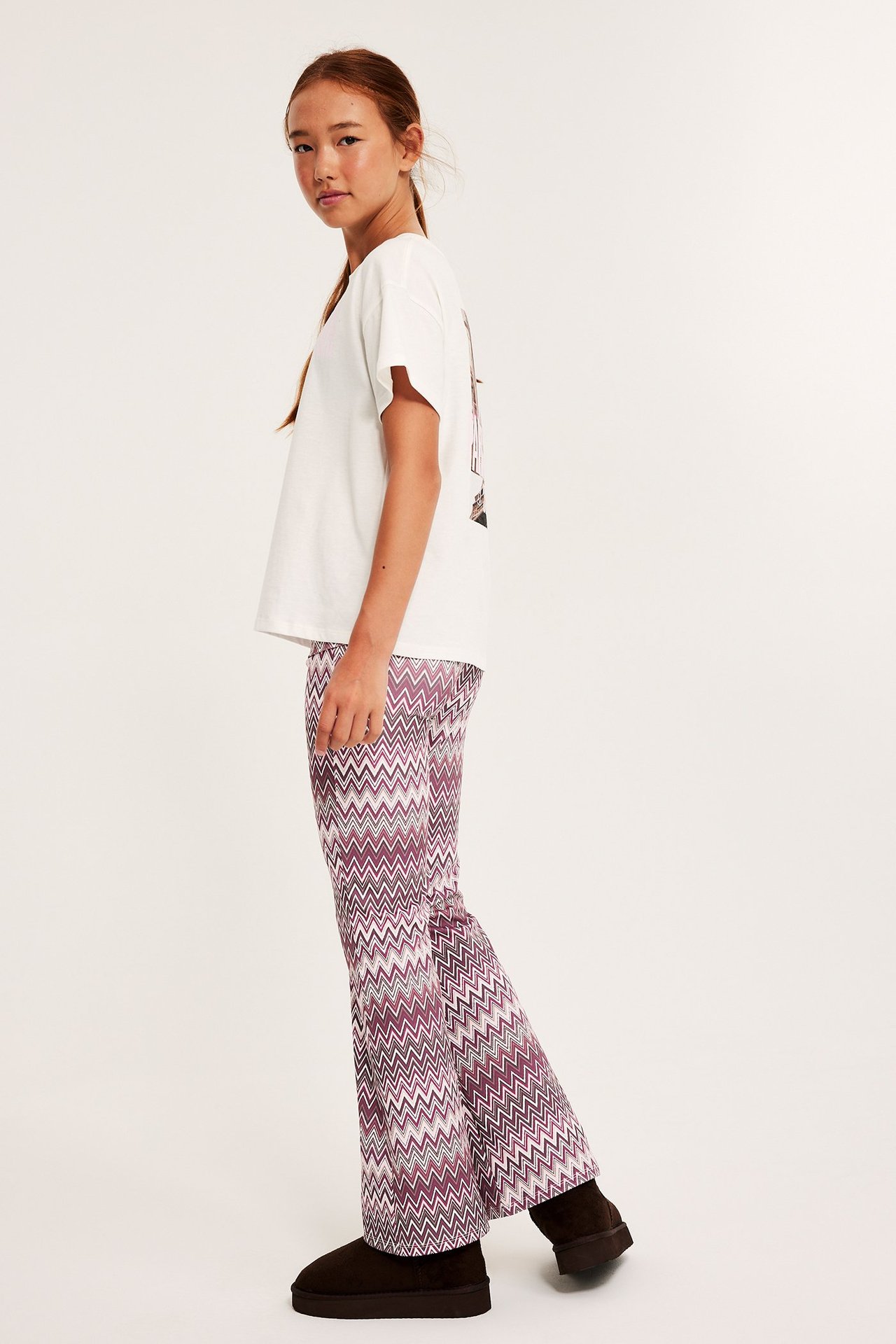 Widok z boku dziecka w ciemnoczerwonych legginsach dzwonach z zygzakiem, białej koszulce i brązowych botkach.