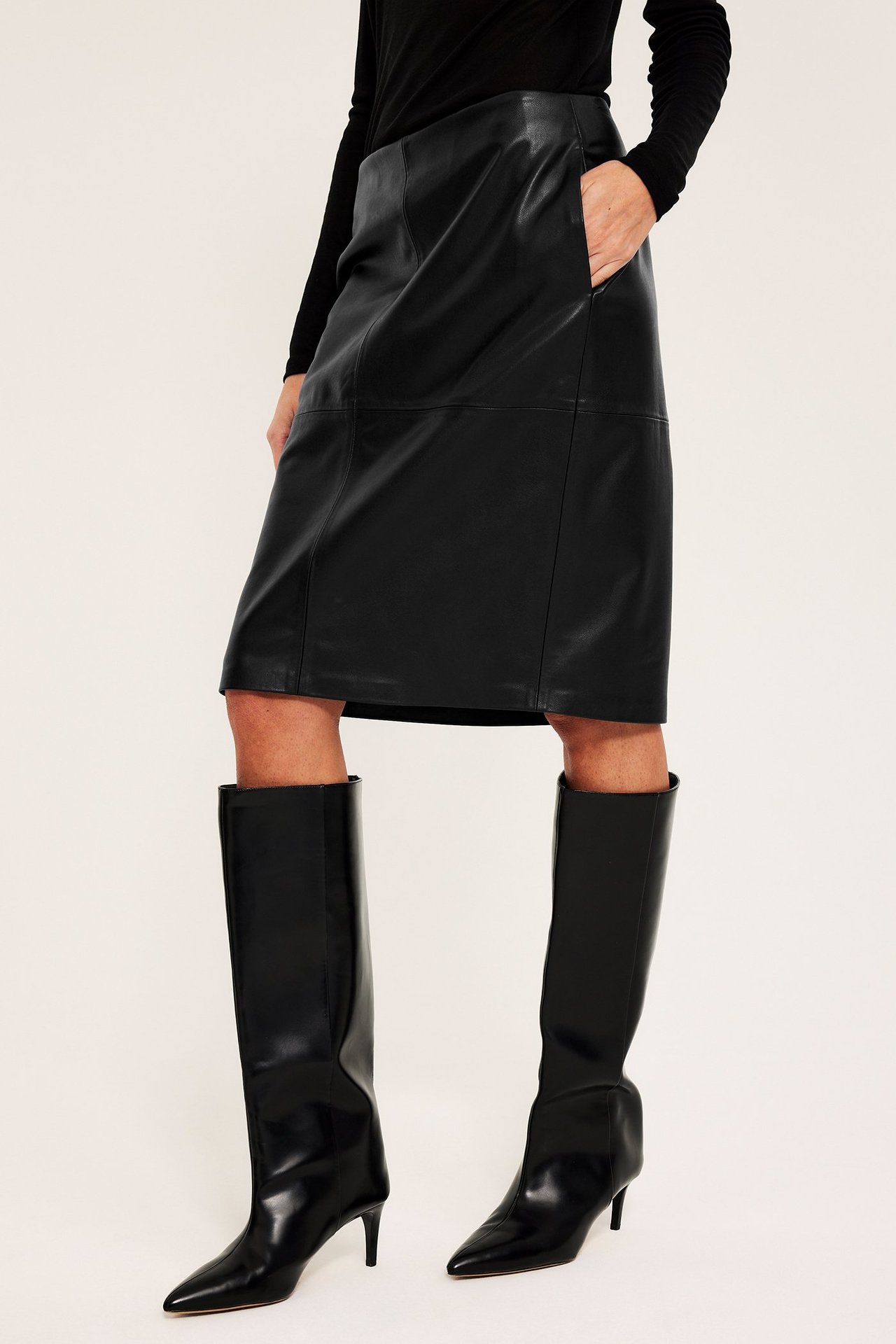 Kvinne i svart blyantskjørt i imitert skinn med sidelommer, svart topp og svarte knelange støvler med hæl.