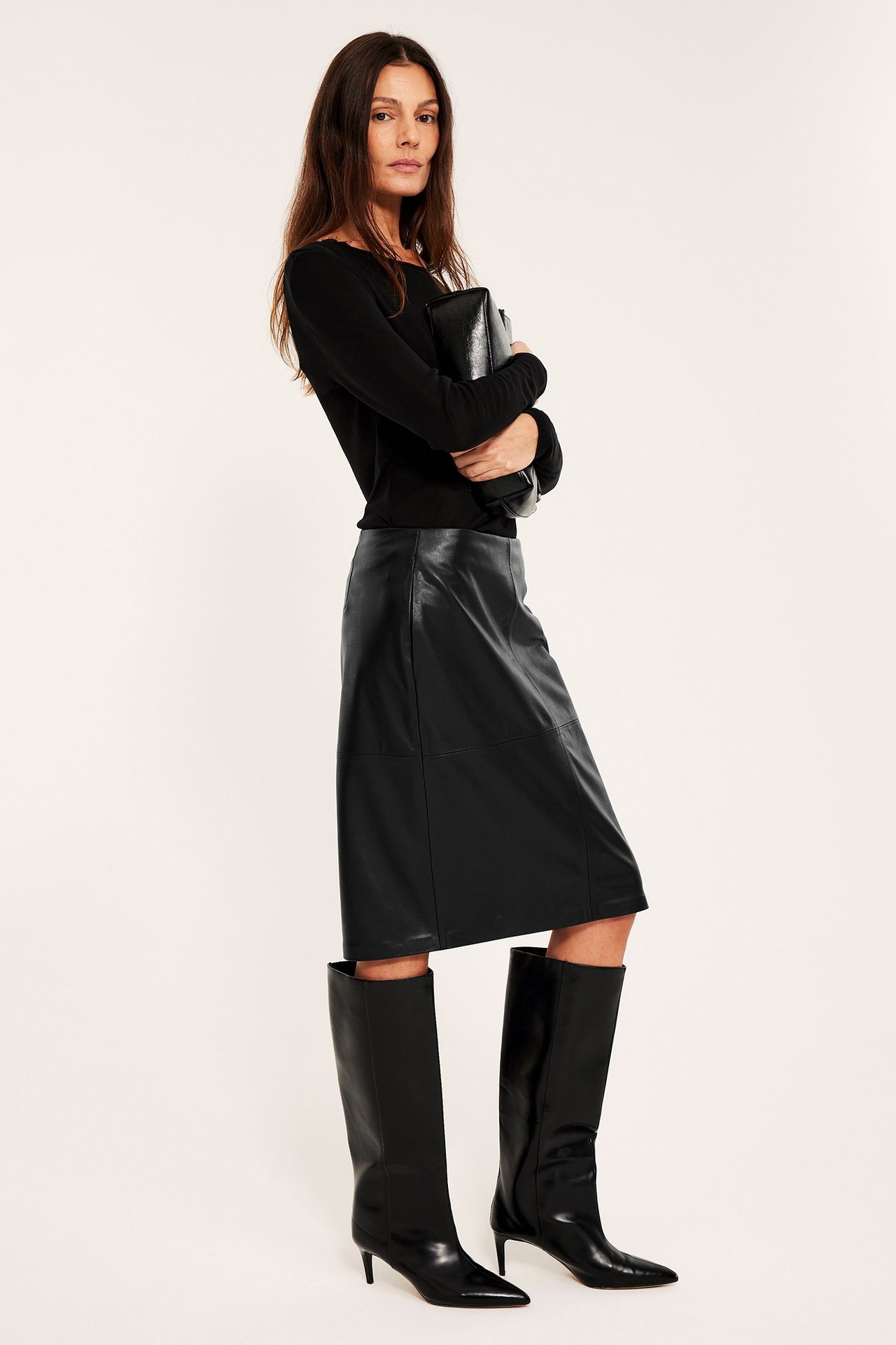 Widok z boku: kobieta w czarnej spódnicy ze skóry ekologicznej, bluzce, kozakach, trzyma kopertówkę.