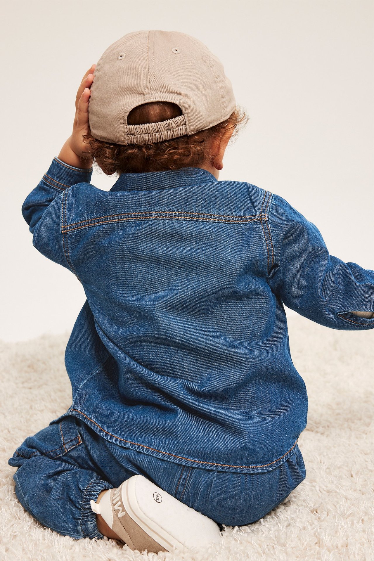 Bakfra: baby i mørk denim bomullsskjorte, matchende jeansbukse, beige caps og hvite sko.