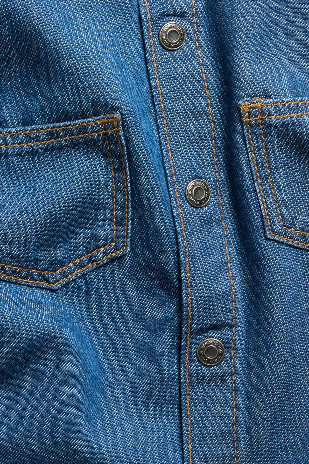 Zbliżenie na ciemnoniebieską koszulę dżinsową z kieszeniami, srebrnymi zatrzaskami i pomarańczowymi przeszyciami.