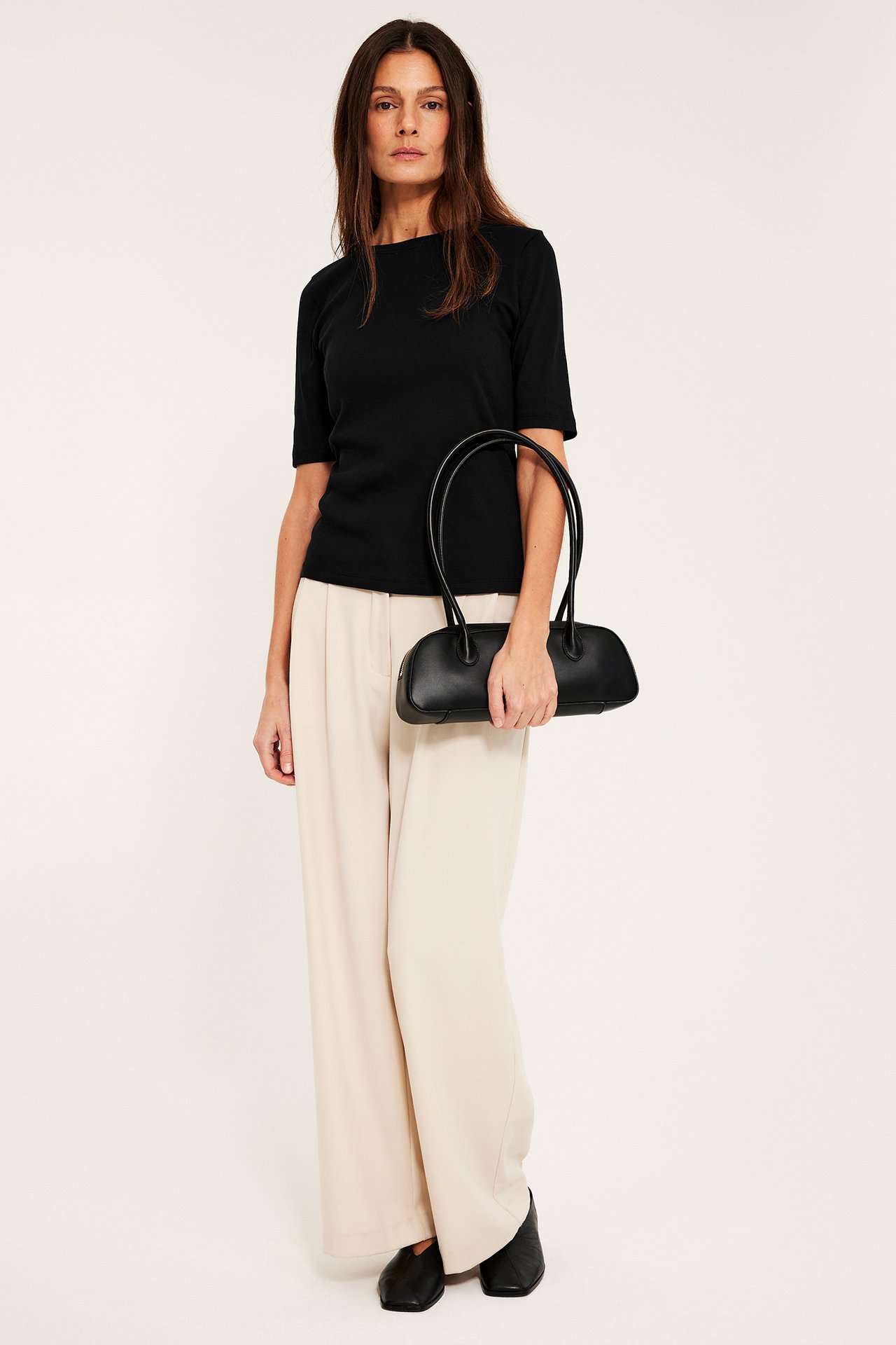 Edestä: nainen mustassa ribbipaidassa, beigenvärisissä leveälahkeisissa housuissa, pitäen mustaa laukkua.