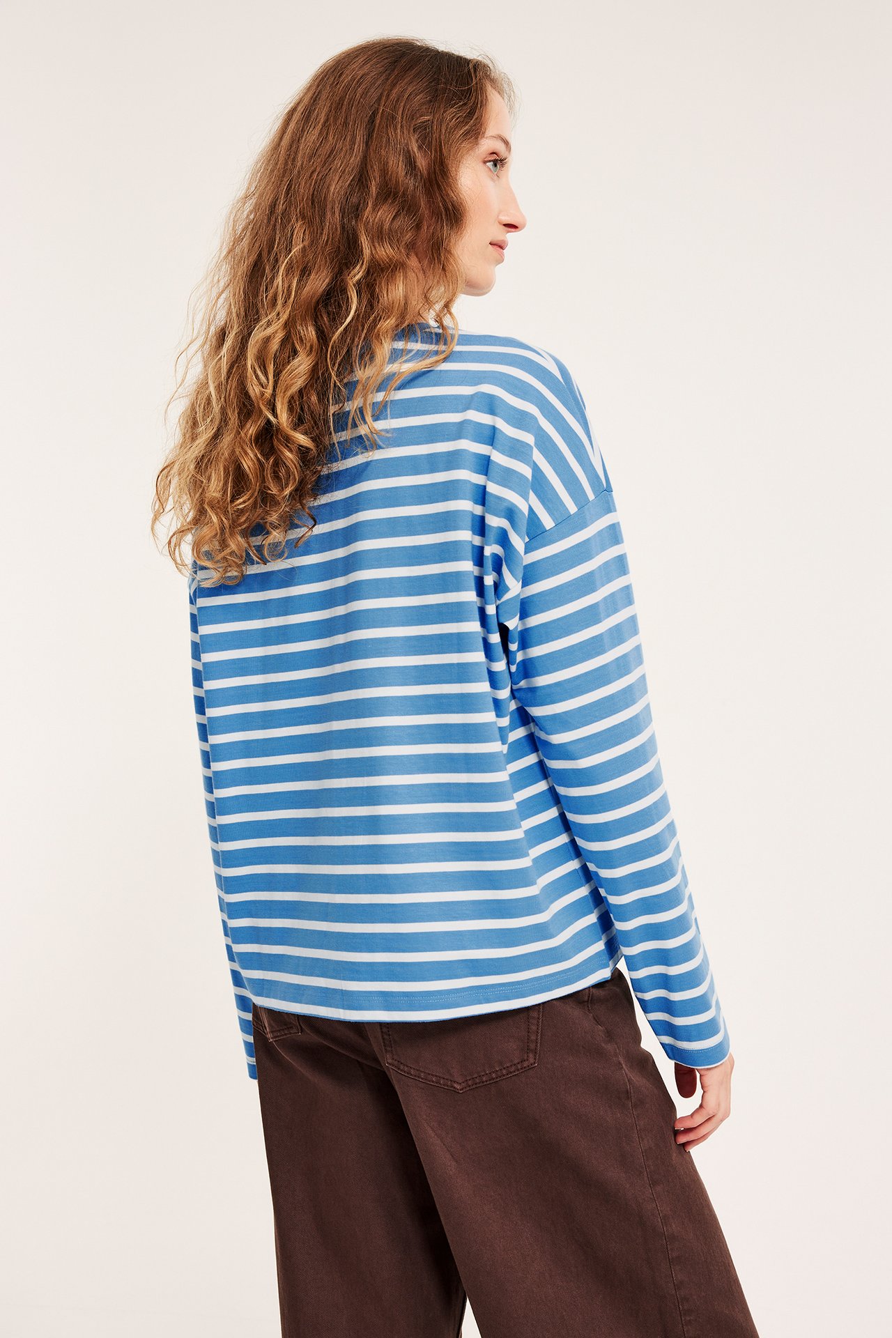 Kobieta w niebiesko-białym pasiastym topie z długim rękawem i brązowych spodniach, widok z tyłu/boku.