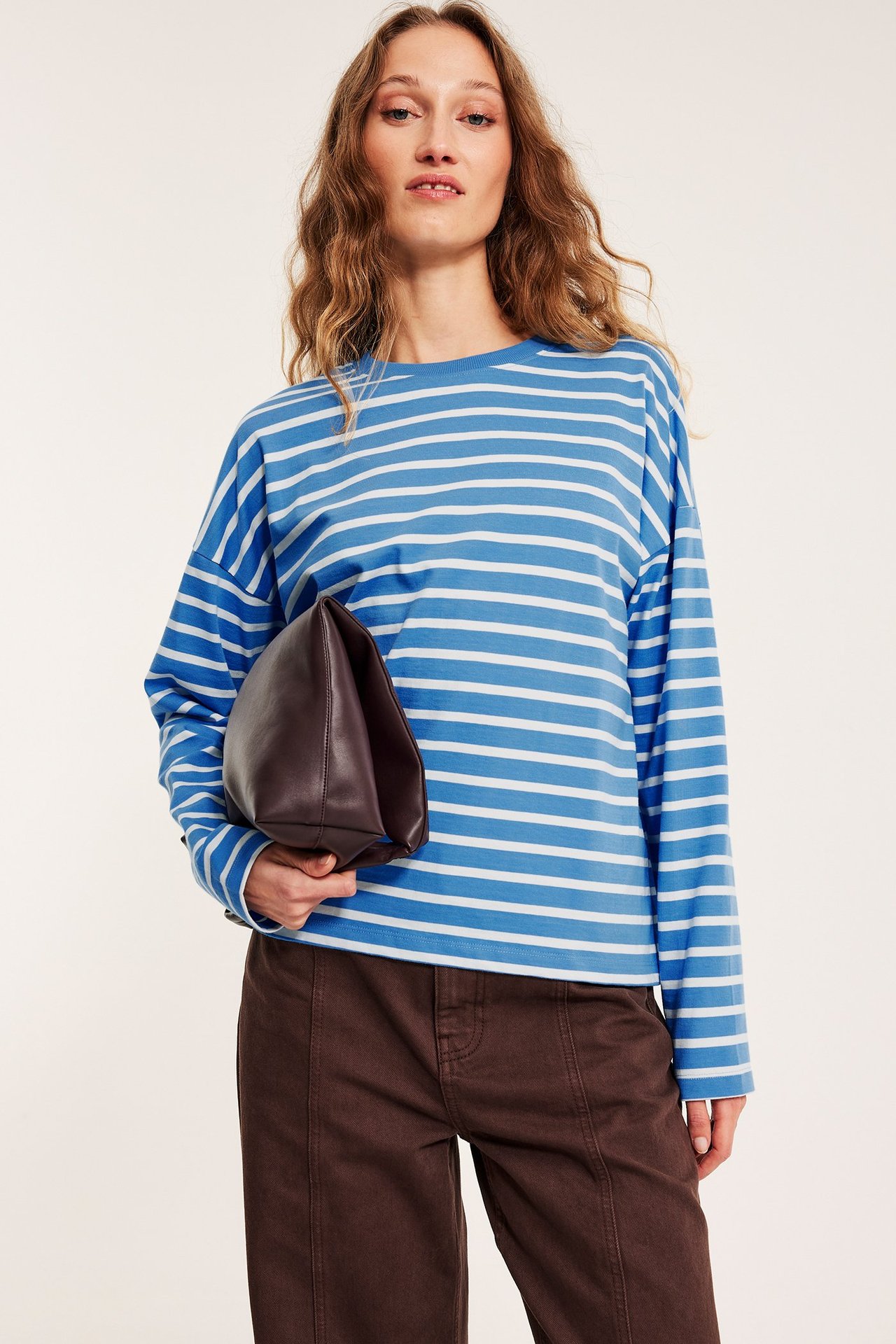 Widok z przodu: Kobieta w niebiesko-białej bluzce w paski z długim rękawem, ciemnobrązowych spodniach, trzyma brązową kopertówkę.