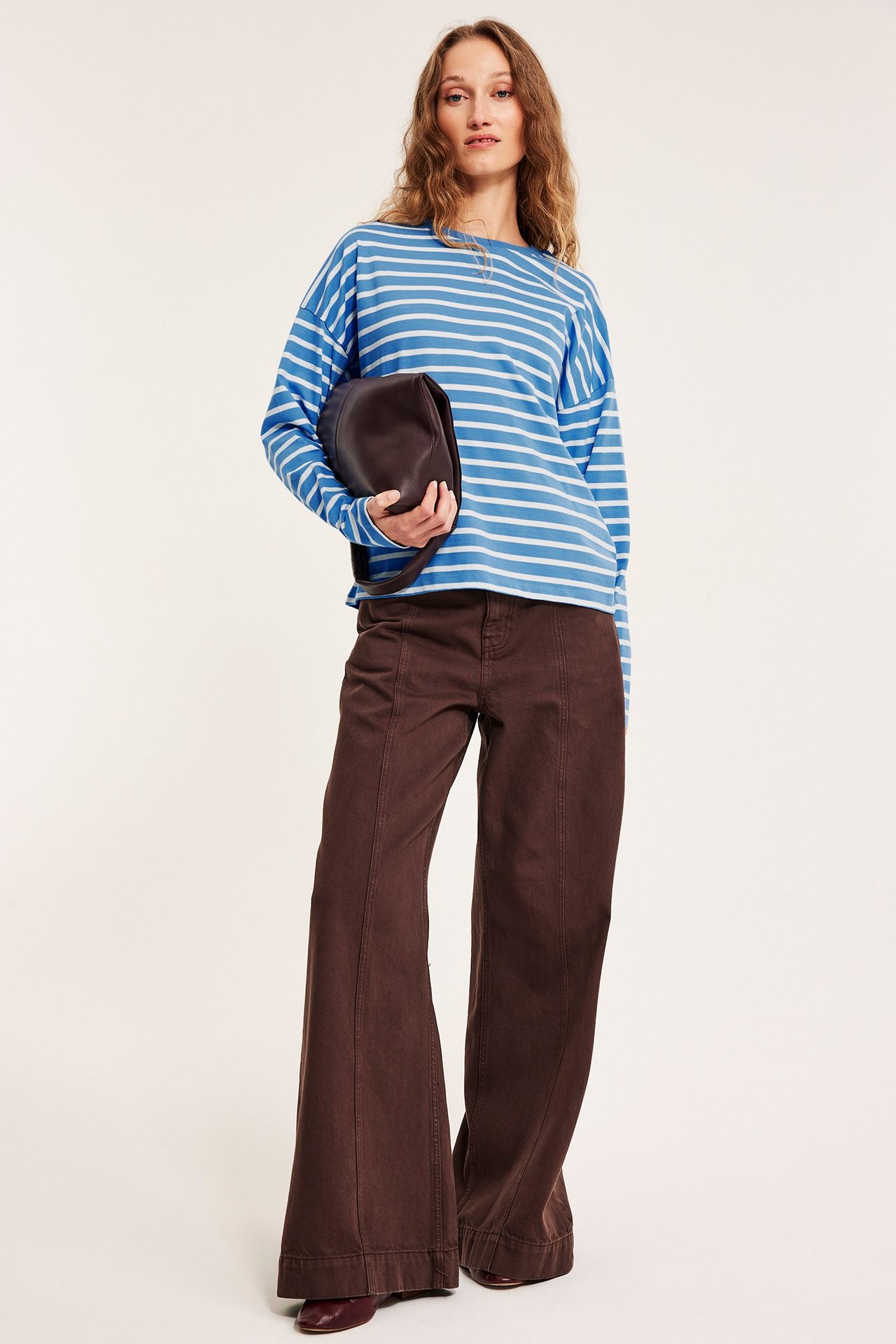 Widok z przodu: Kobieta w niebieskiej bluzce w paski, brązowych szerokich spodniach, trzyma ciemnobrązową torebkę.