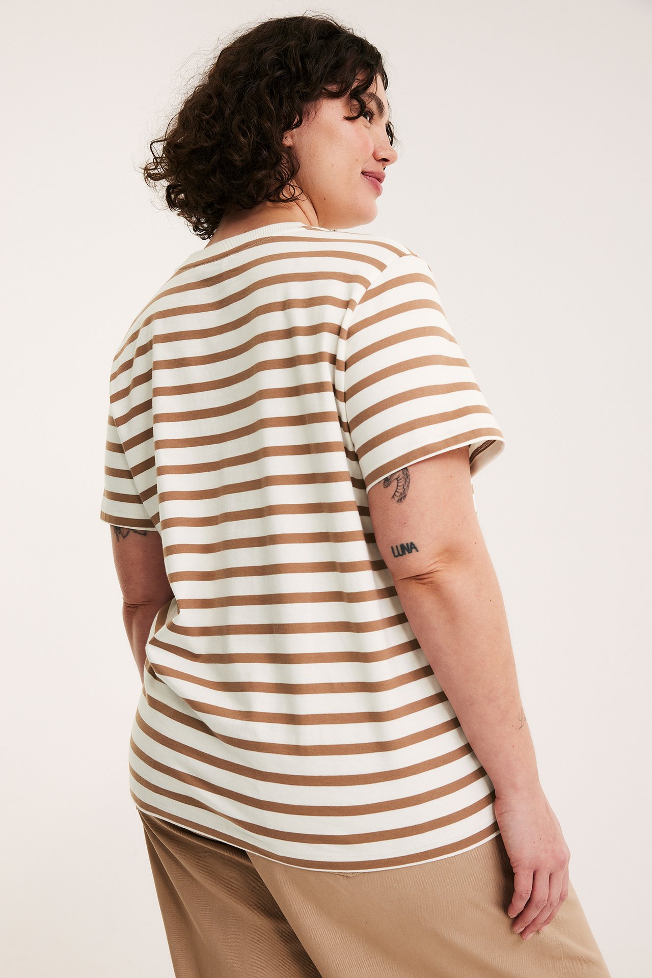 Bak-/sidovy av en kvinna som bär en beige- och vitrandig kortärmad bomullstopp och beiga byxor.