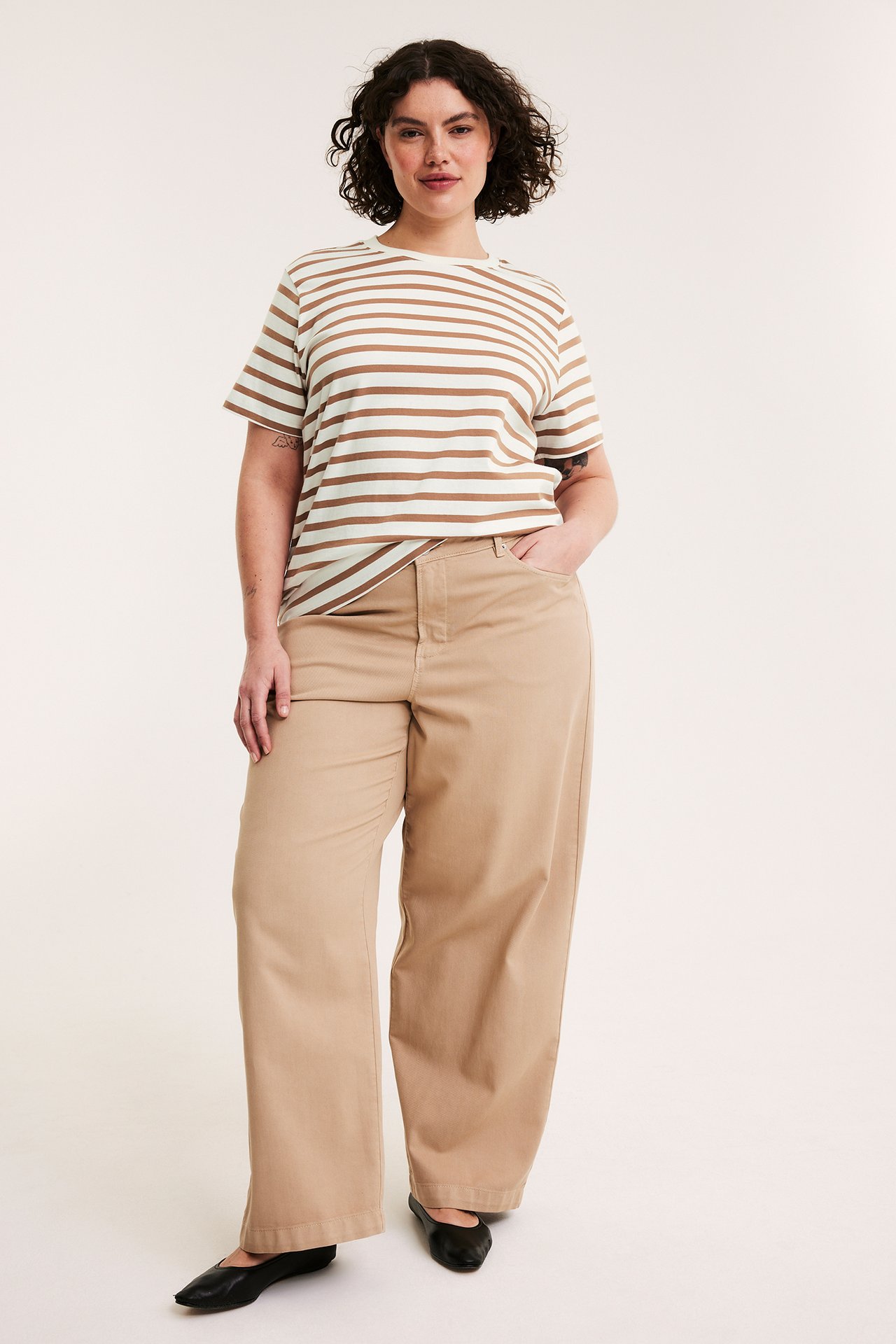 Edestä kuvattu nainen beigessä ja valkoisessa raidallisessa lyhythihaisessa topissa, beigessä leveälahkeisissa housuissa, mustissa kengissä.