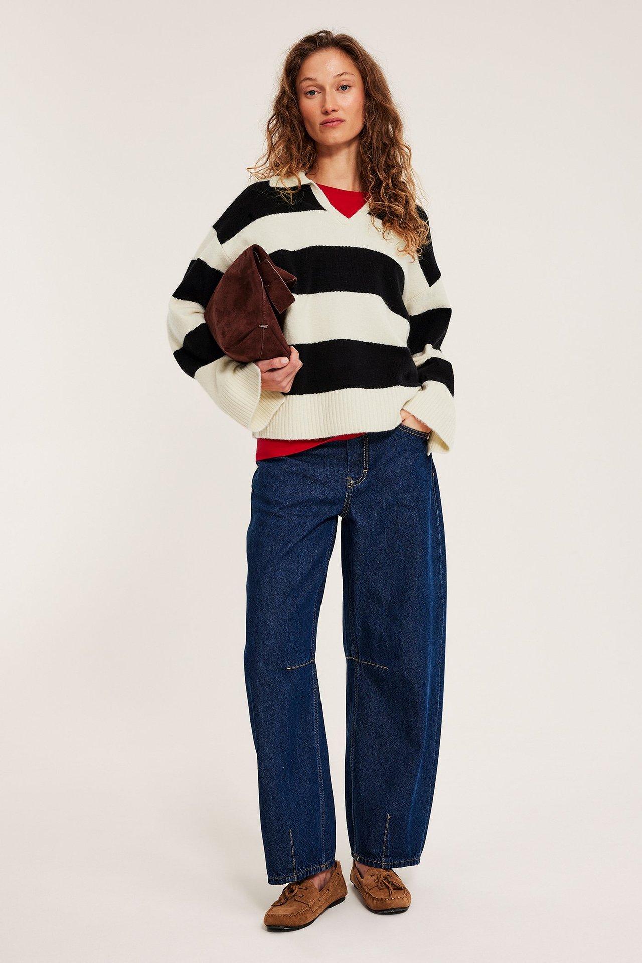 Kvinne i offwhite-svart stripet strikkegenser, rød topp, blå jeans, brune mokasiner, holder brun clutch.