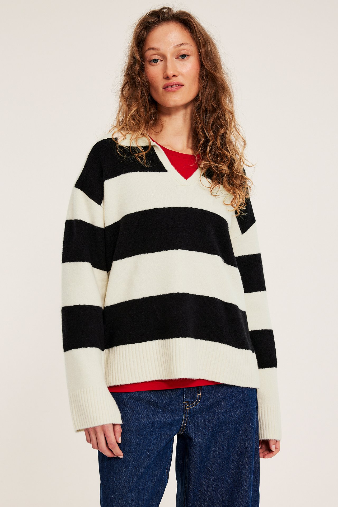 Forfra, kvinne i offwhite-svart stripete V-hals genser over rød t-skjorte og blå jeans.