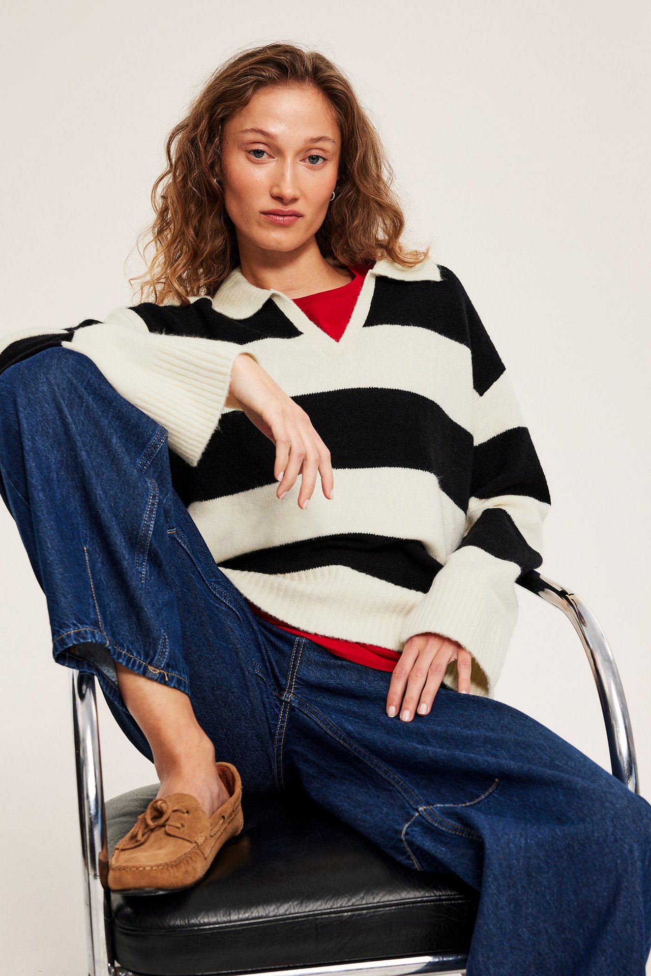 Forfra: kvinne i svart- og offwhite-stripet strikket genser, rød topp, blå jeans, brune loafers.