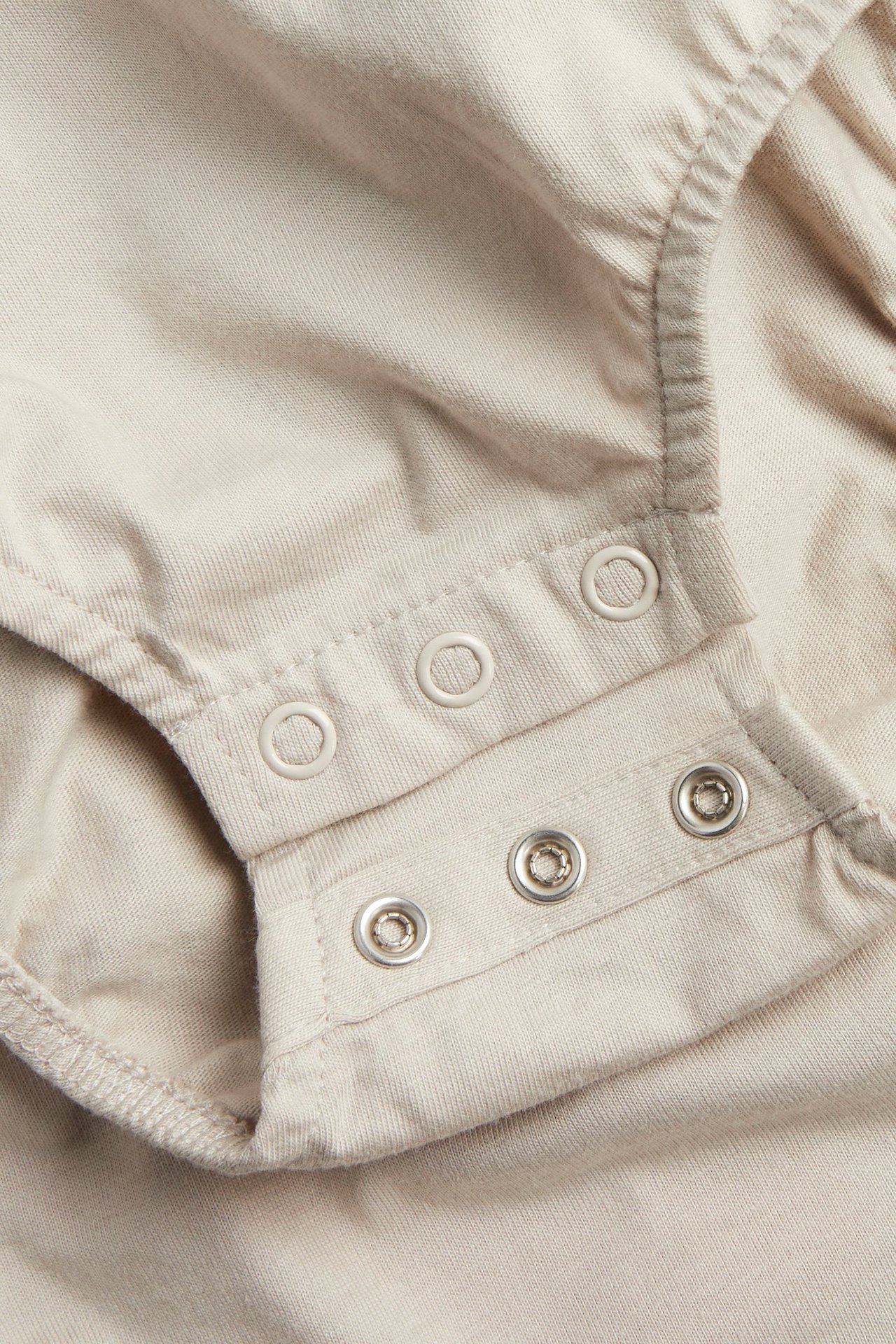 Närbild av en beige babybody i bomull med tryckknappar vid gren och axel.