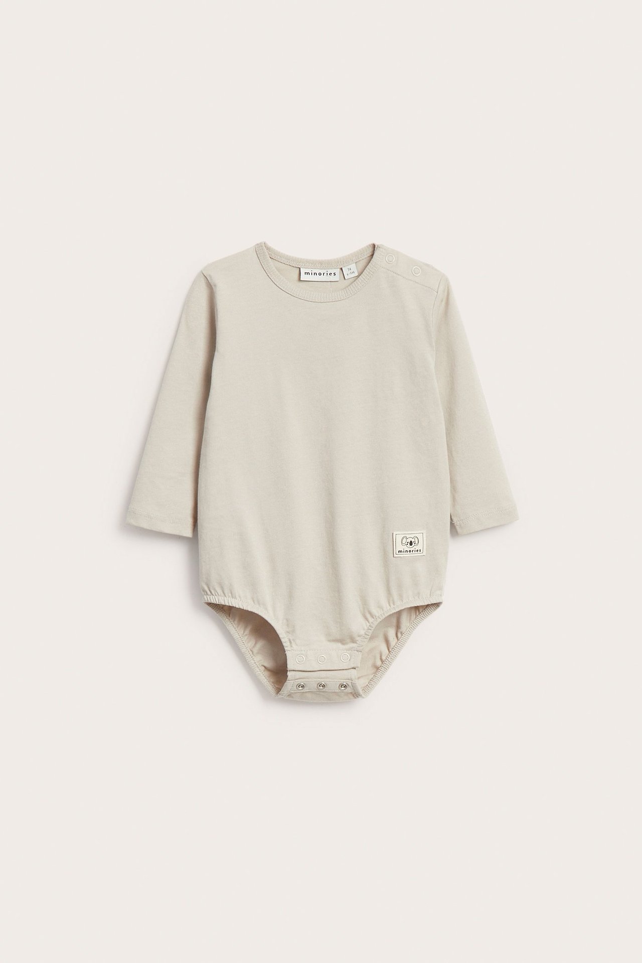 Front view: oversized beige bomullsbody med lång ärm för baby, med vävt märke och tryckknappar.