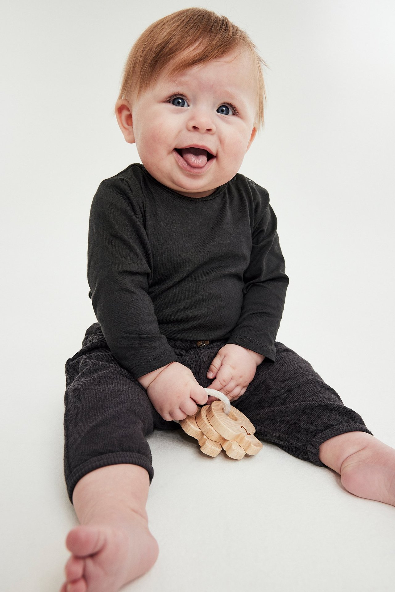 Edestä vauva mustassa pitkähihaisessa puuvillabodyssa ja tummissa housuissa, pitäen puulelua.
