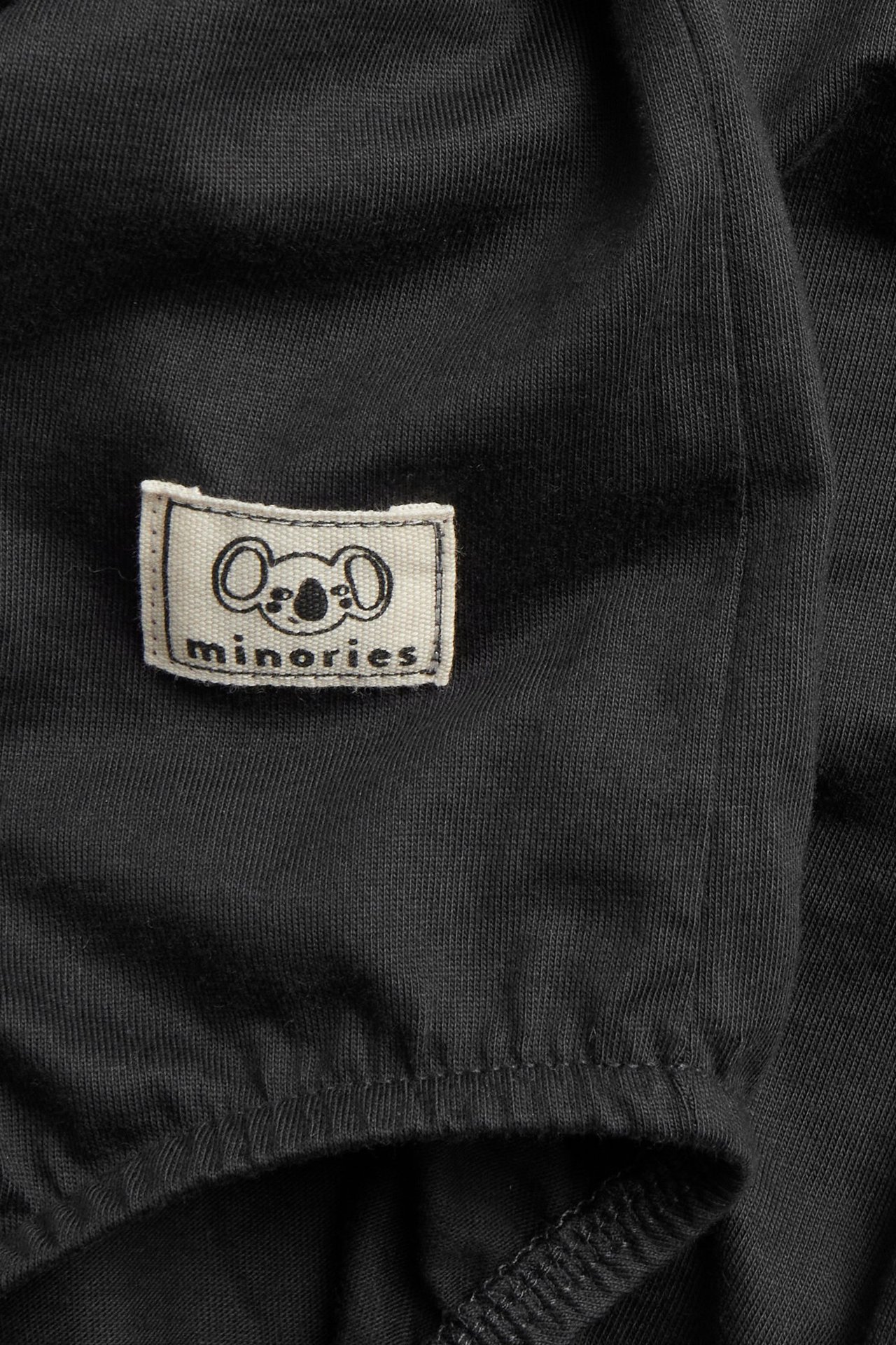 Zbliżenie na czarne, bawełniane body niemowlęce z tkaną naszywką minories z koalą.